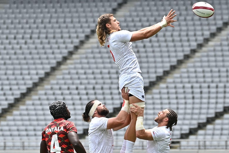 Two Team USA men's rugby sevens players lift their teammate in the air, while a Team Kenya player looks on. The player in the air has a long blond mullet, and his arms extended to catch the ball.