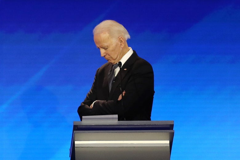 Joe Biden crosses his arms across his chest and looks down while behind a podium onstage.