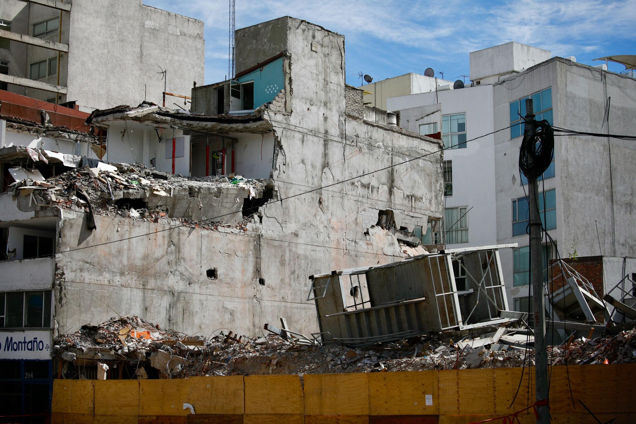 A badly damaged, grey brick building is seen next to a space left by a building that collapsed, leaving piles of rubble.  