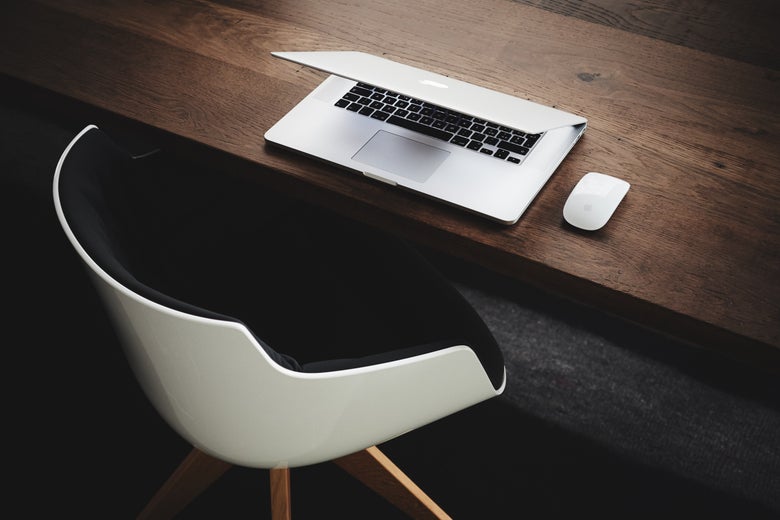 A laptop sits half-open on a wooden desk in front of an empty chair.