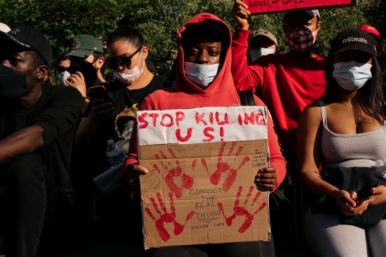 A man in a red hoodie and face mask holds a sign with handprints that says "Stop Killing Us." He is standing in front of other protesters wearing face masks and carrying signs.