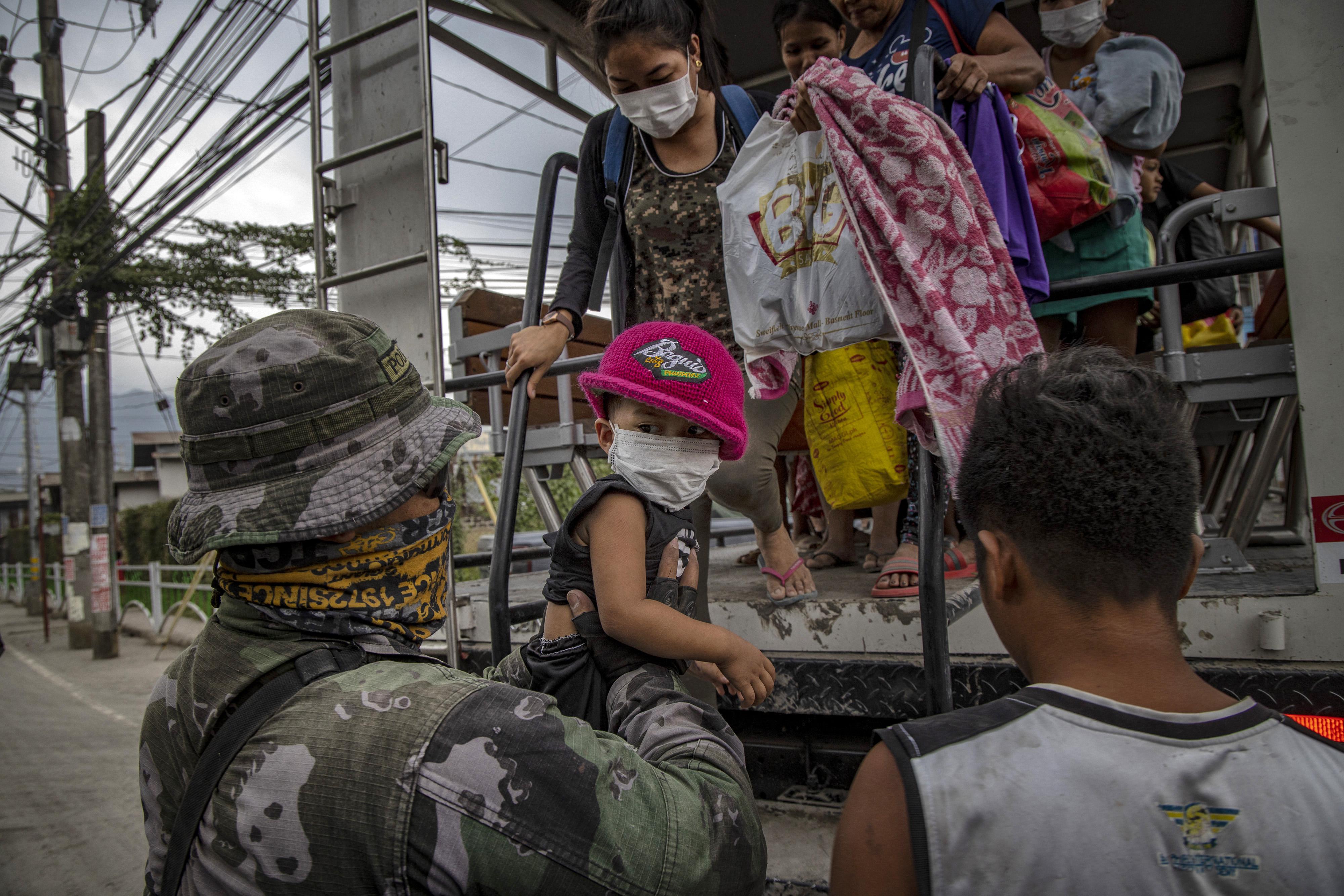A man in a military uniform holds a child wearing a mask as adults carrying a small number of possessions prepare to step down from a vehicle.