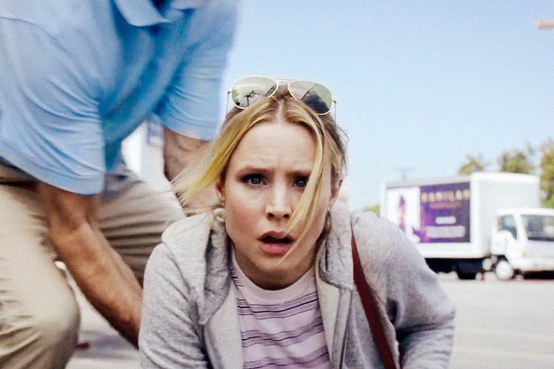 In a scene from The Good Place, Eleanor Shellstrop, played by Kristen Bell, looks dazed as she gets to her feet in a parking lot. A man in a blue polo helps her up, and a truck can be seen in the background.