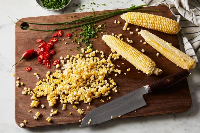Corn on a cob with some of it shucked, chopped red pepper, and chopped scallions on a wooden cutting board.