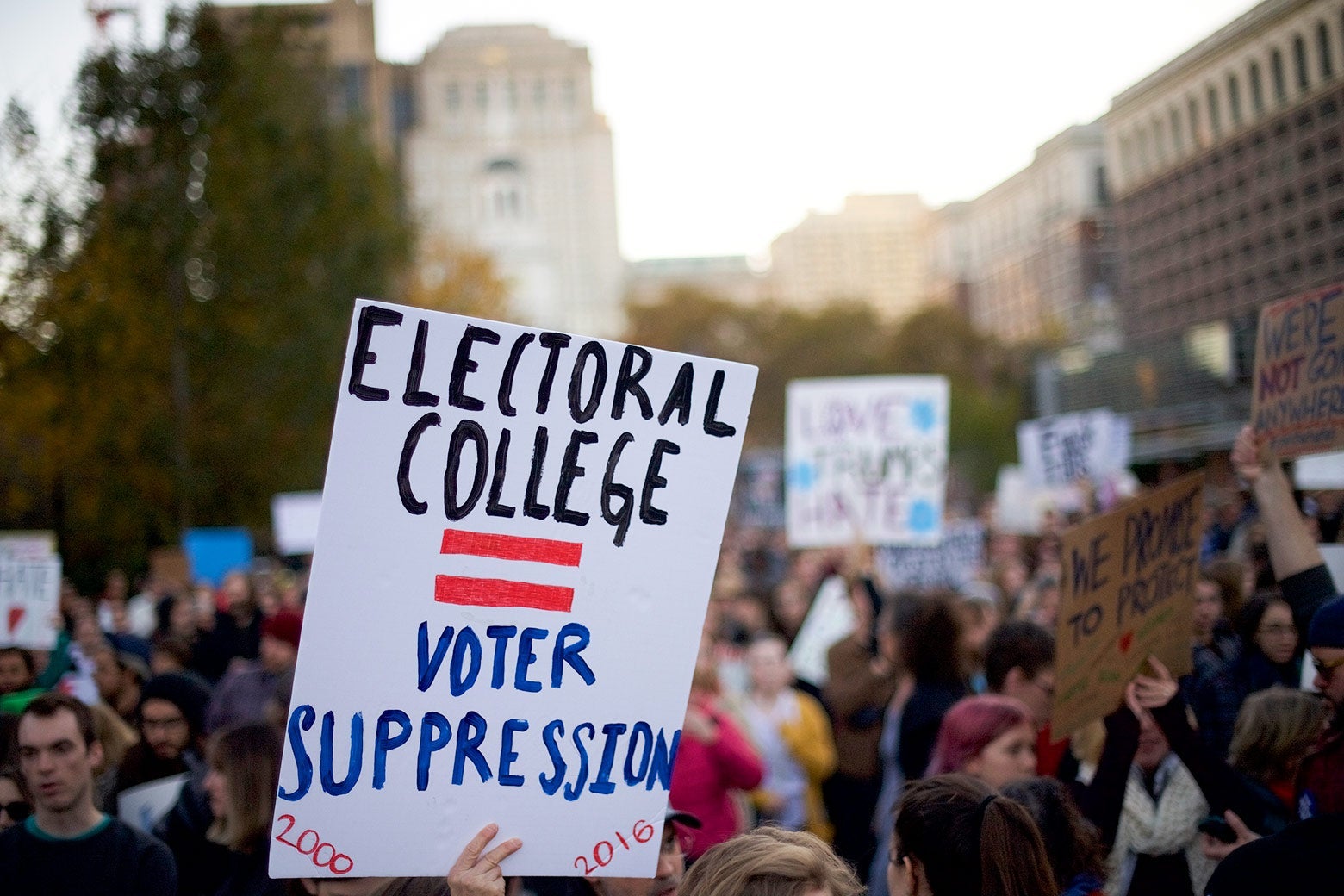 A protester in a crowd holds up a sign that says "Electoral College = Voter Suppression."