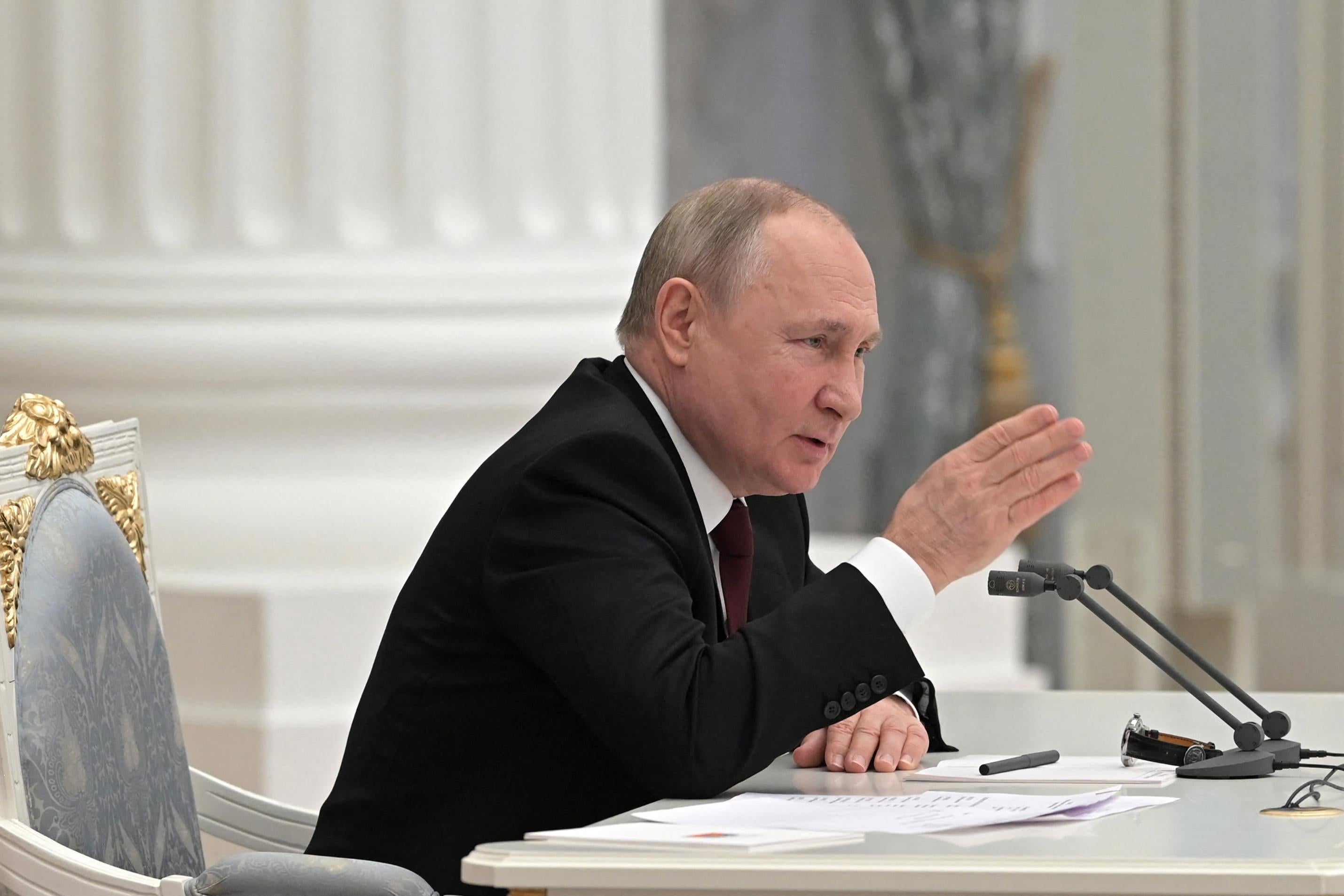 Vladimir Putin makes a chopping motion with his hand as he sits at a table in front of microphones.