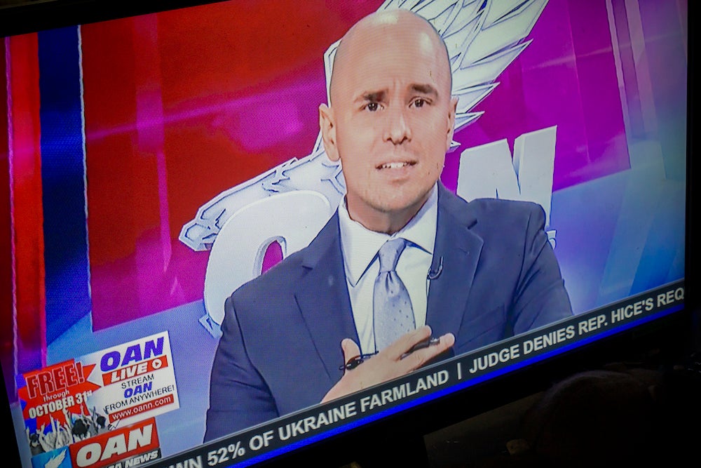 OAN host Pearson Sharp, a bald man in a grey suit, appearing on TV.