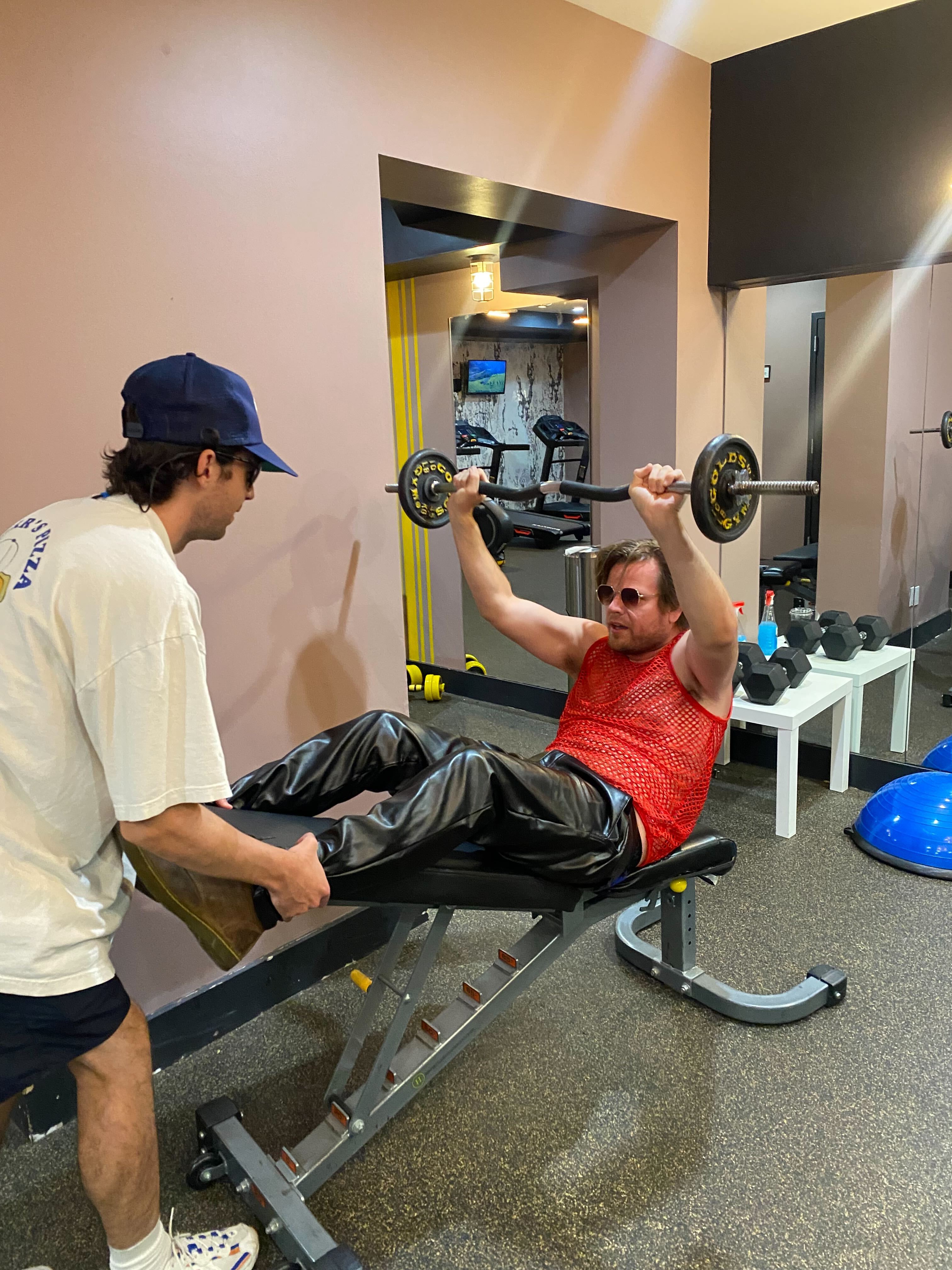 A man wearing sunglasses and a mesh tank top lifts on a barbell on an inclined weight bench.