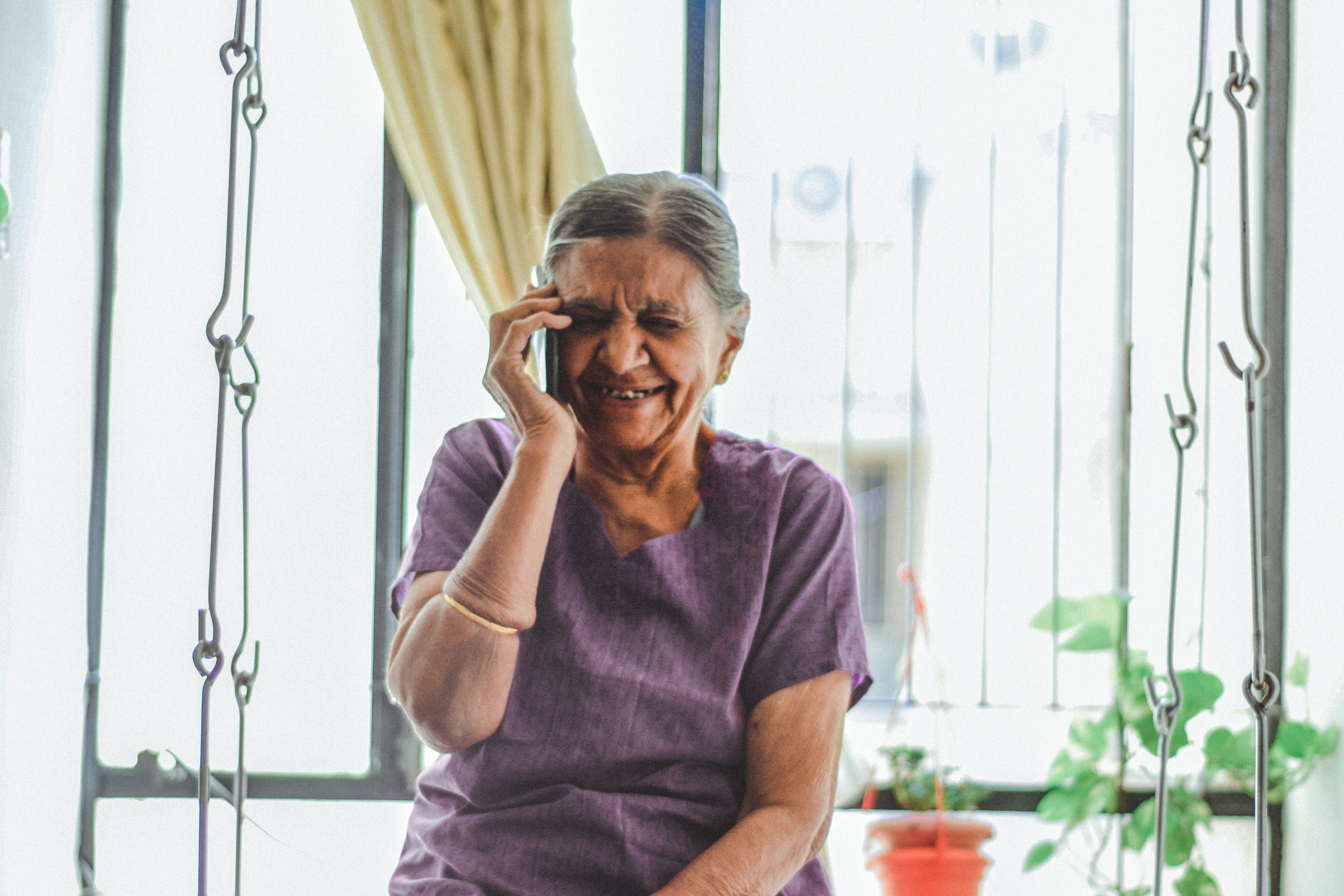 An elderly woman listens on a cellphone in front of a window.