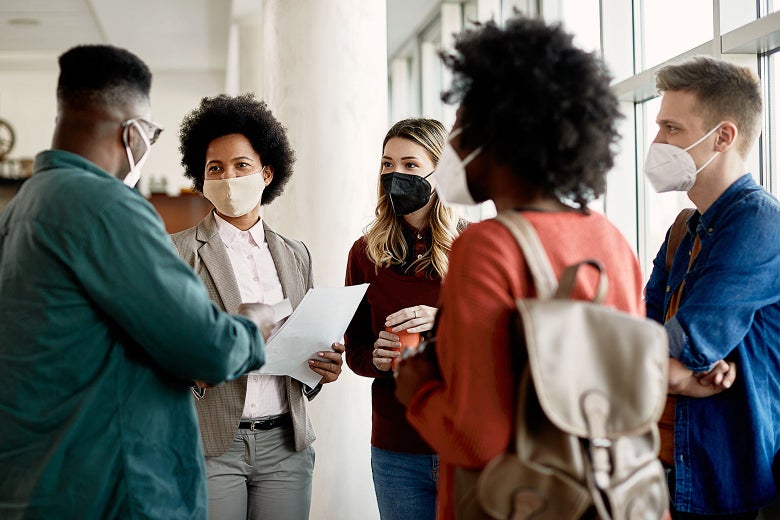 A group of five people in face masks stand in a circle inside a student hall.