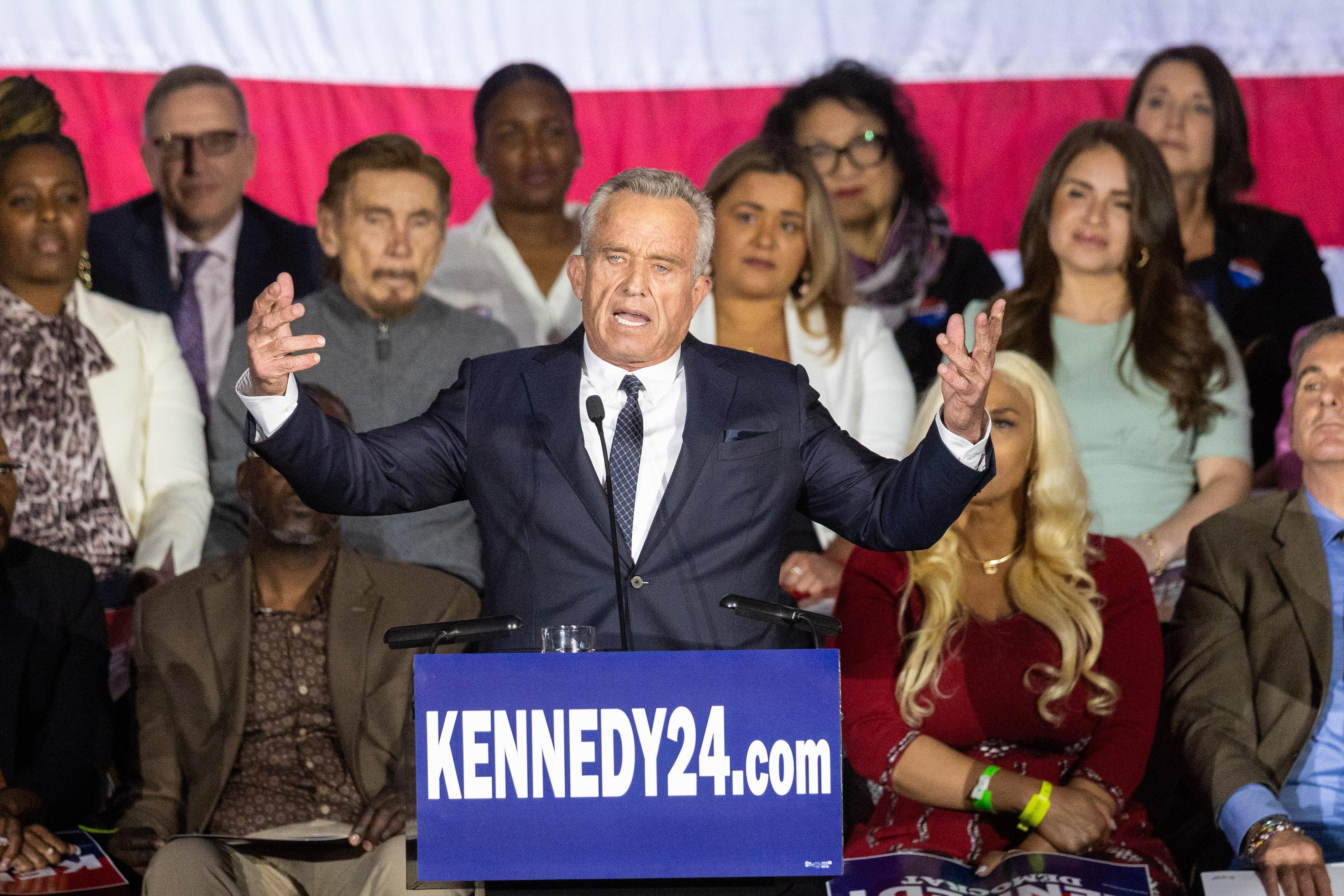 A man behind a podium with a sign that says "Kennedy 2024" stands before a large crowd with his hands raised. 