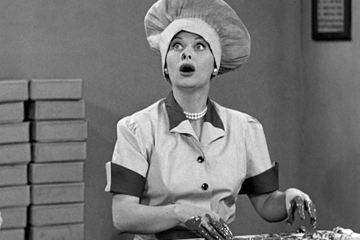 A black and white photo. Lucille Ball, wearing a large chef's hat, sits open-mouthed and wide-eyed before a conveyer belt. Her fingers are covered in chocolate.