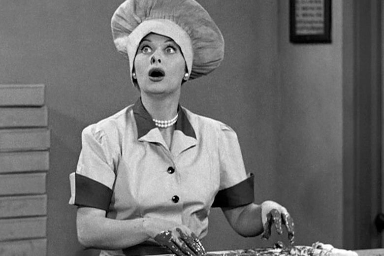 A black and white photo. Lucille Ball, wearing a large chef's hat, sits open-mouthed and wide-eyed before a conveyer belt. Her fingers are covered in chocolate.