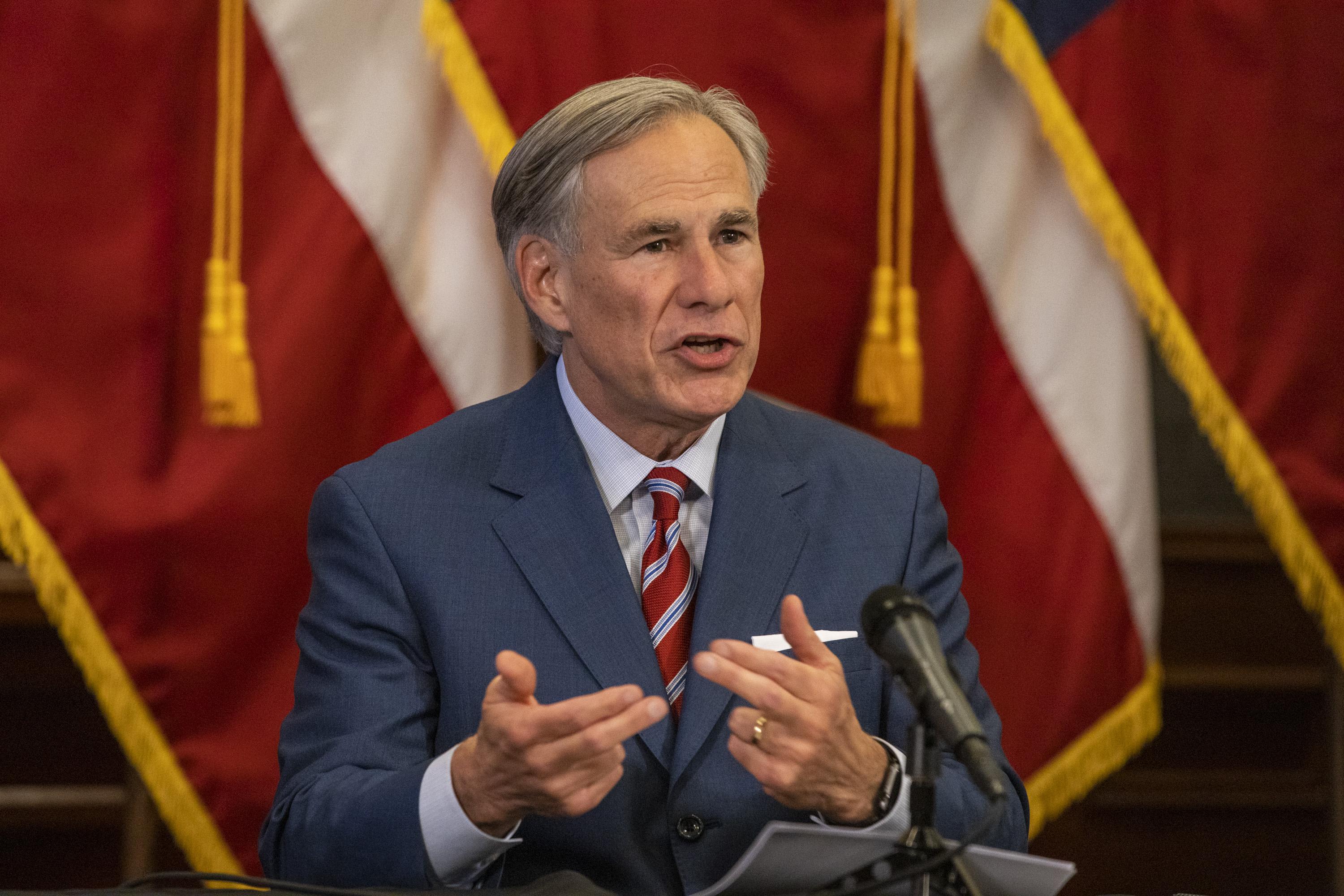 Texas Gov. Greg Abbott speaks in front of American flags, gesturing with his hands.