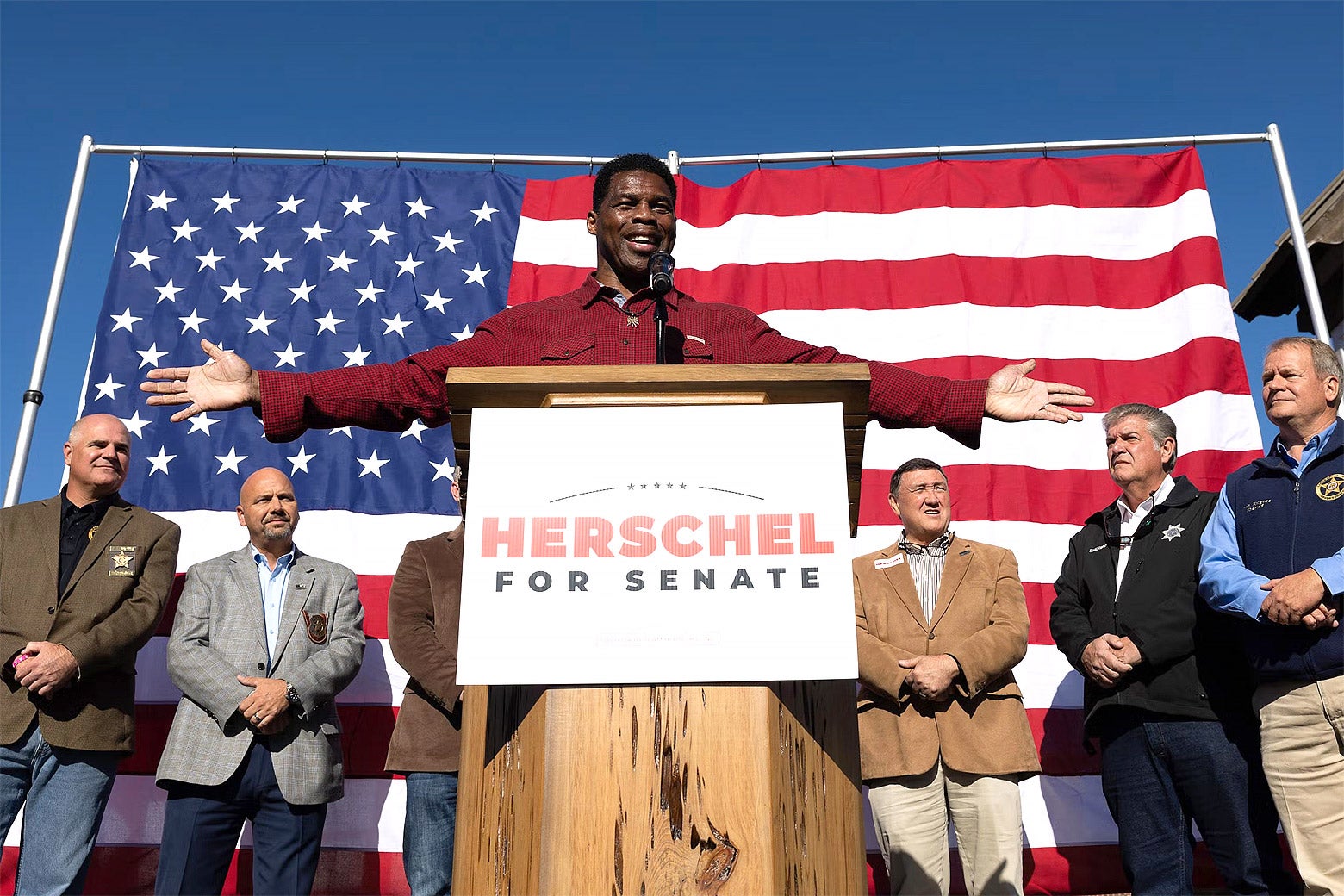 Georgia Republican Senate nominee Herschel Walker addresses the crowd during a campaign stop on Oct. 20 in Macon, Georgia. He has his arms wide open in front of an American flag surrounded by several old white men.