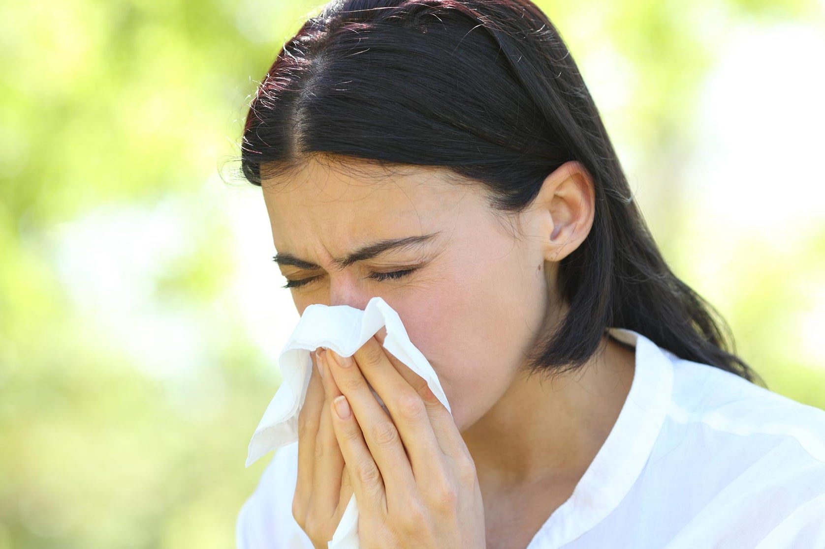 A woman sitting outside sneezes into a tissue.