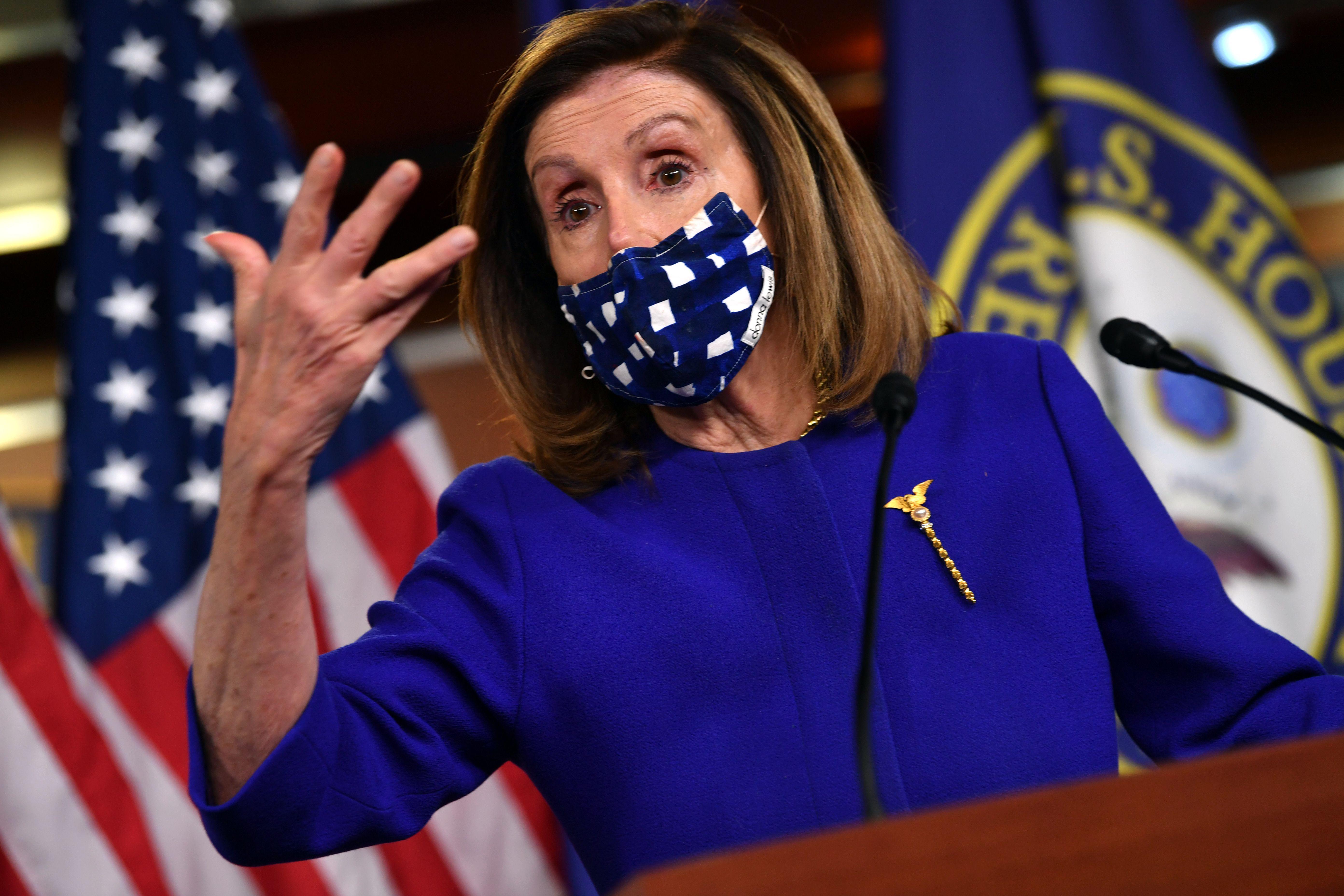 Nancy Pelosi gesticulates while wearing a mask and standing at a podium in front of a U.S. flag and a House of Representatives flag.