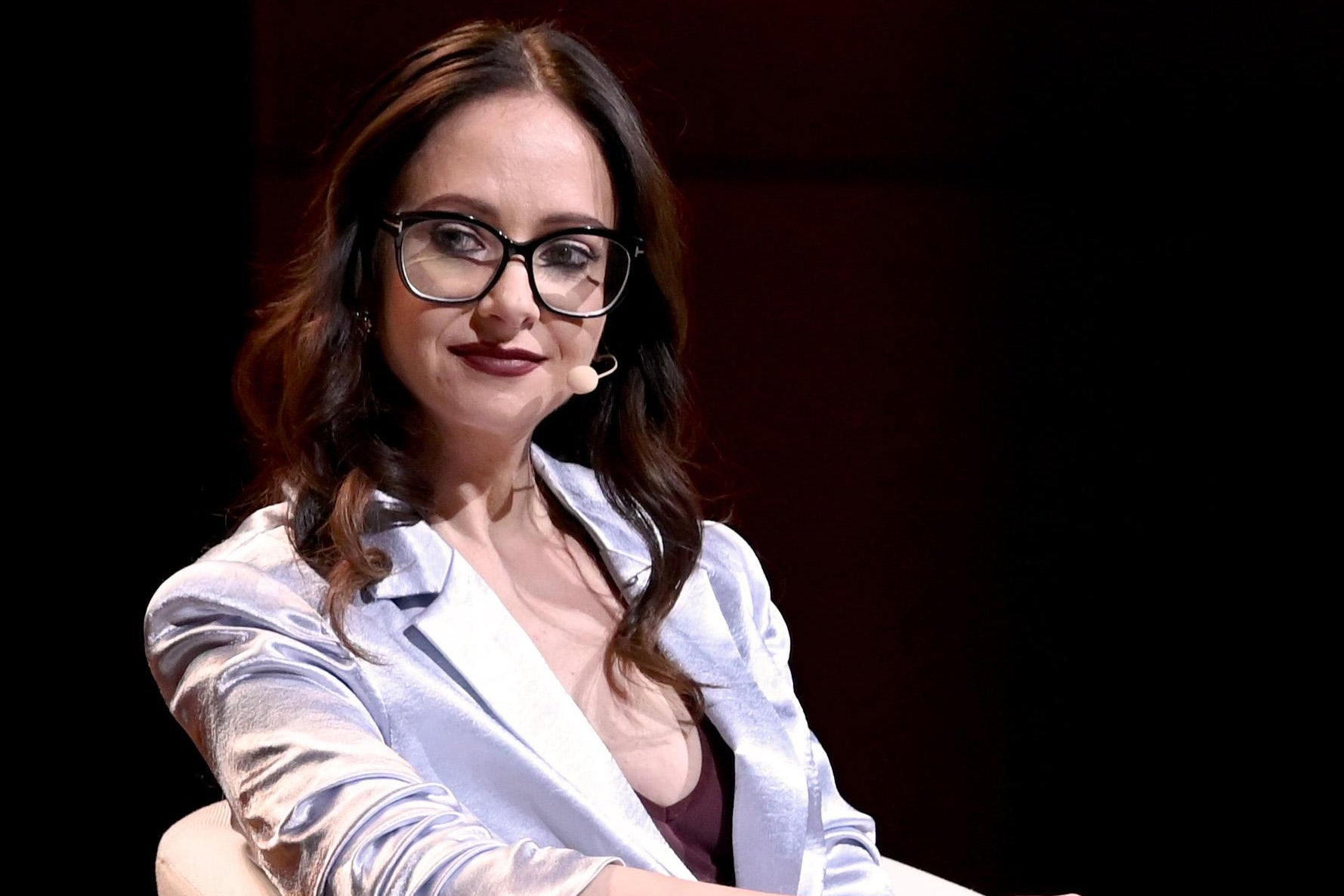 A woman with brown hair, glasses, and a hands-free microphone sits on a stage in a chair.