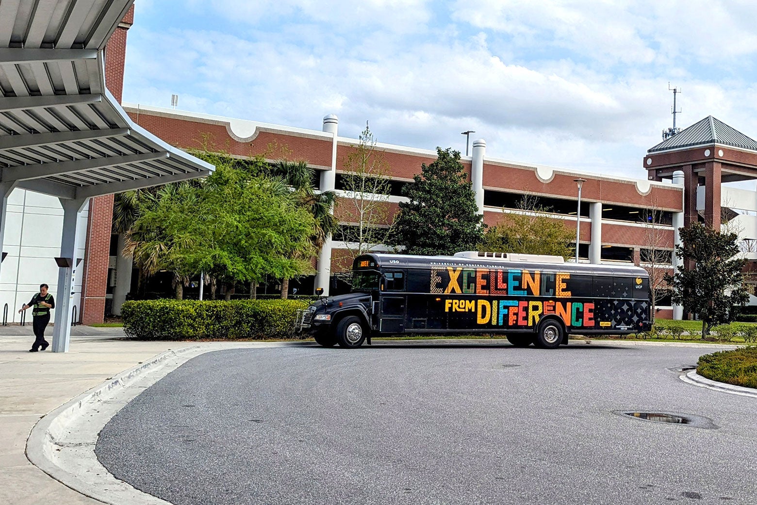 A black bus parked outside a red-brick college building reads "Excellence From Difference" in colorful letters.