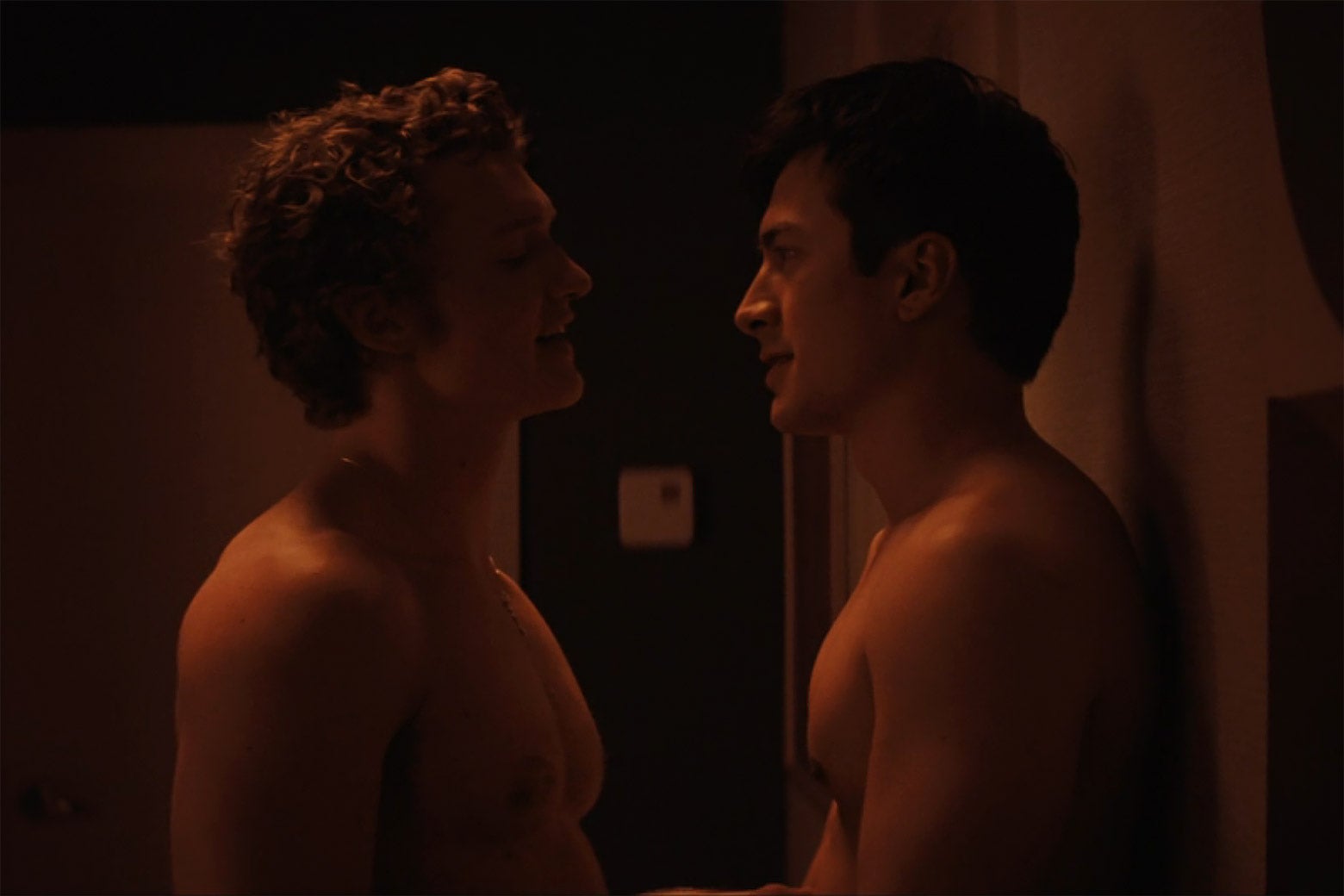 Two shirtless men stare at each other in a dimly lit hotel room.