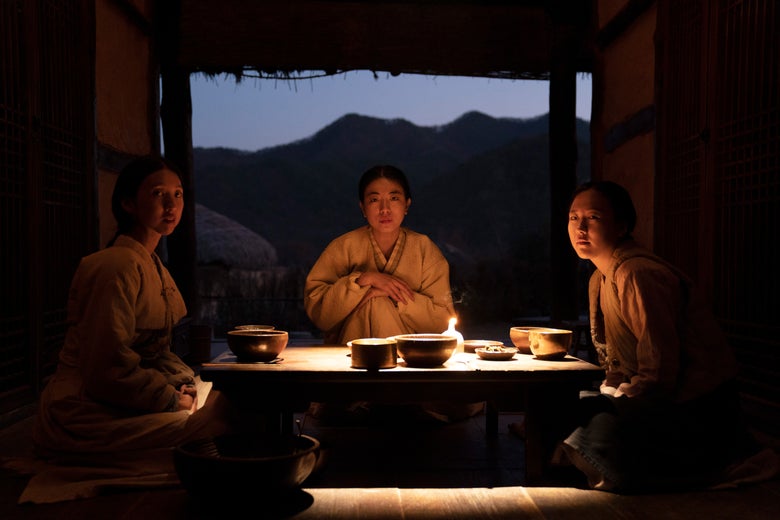 Three Korean women in plain traditional dress sit at a low table with bowls of food in front of them.