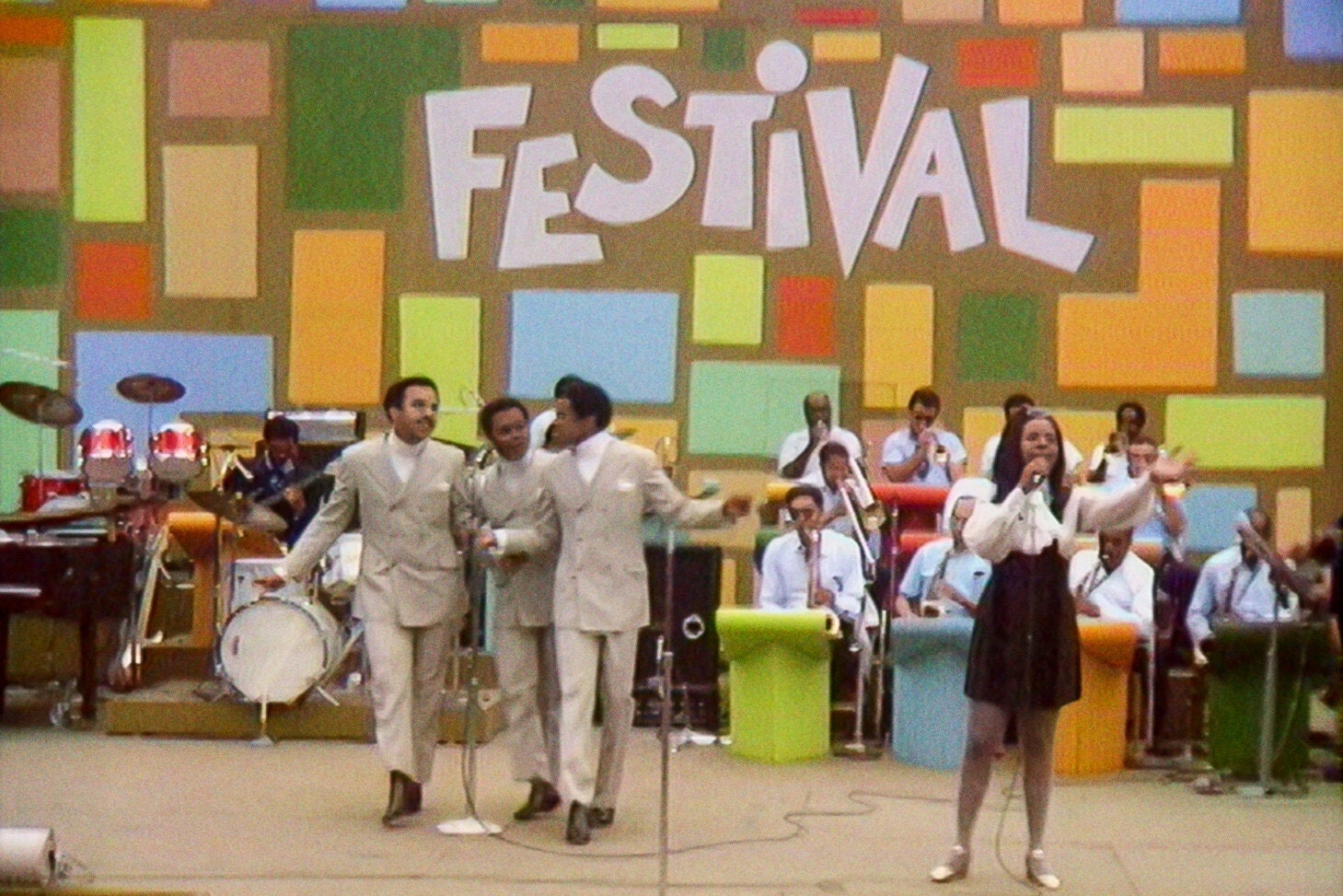 Four artists (three men in suits, one woman in a dress) stand on a stage performing against a multicolored backdrop that reads FESTIVAL.