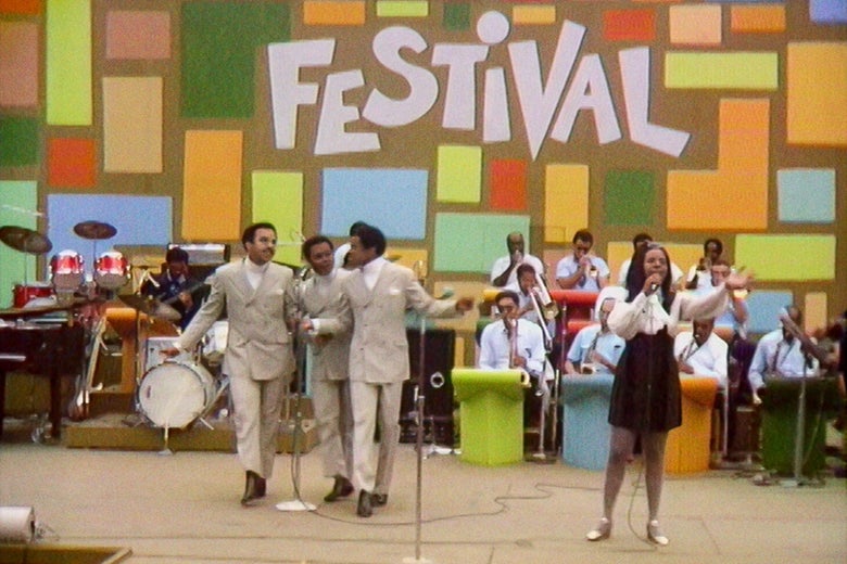 Four artists (three men in suits, one woman in a dress) stand on a stage performing against a multicolored backdrop that reads FESTIVAL.