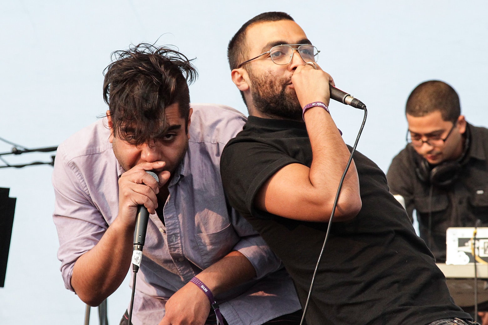 The two rappers lean on each other holding microphones on stage outdoors. 
