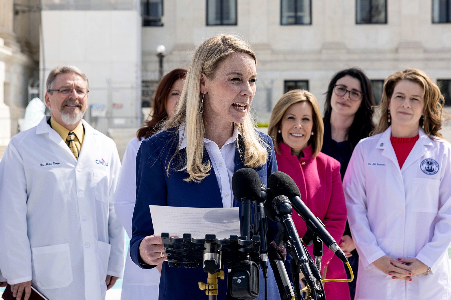 Hawley reads from a piece of paper at a lectern with doctors in lab coats behind her.