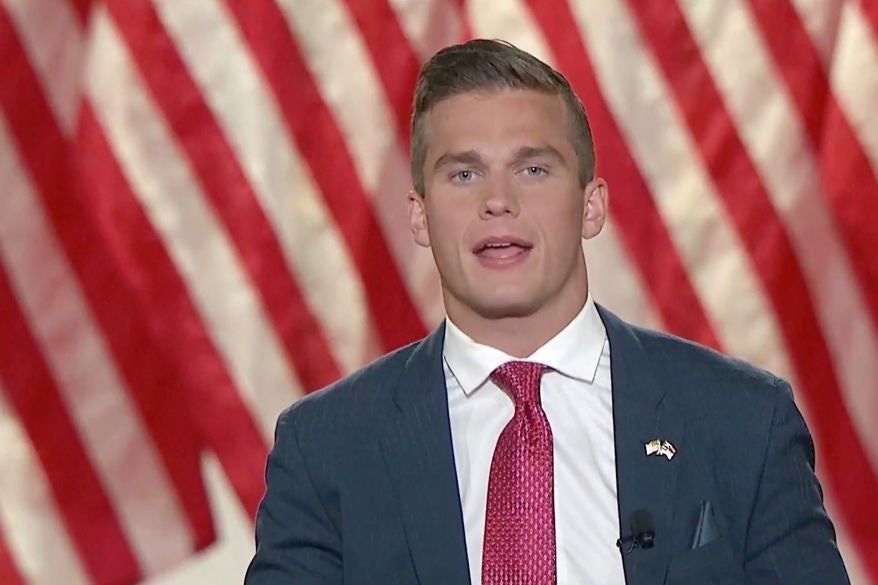 Cawthorn, a young man with a firm jawline and blond hair, speaks against a backdrop of American flags while wearing a blue-gray suit jacket with a pink tie and an American flag pin.