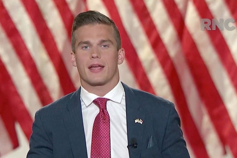 Cawthorn, a young man with a firm jawline and blond hair, speaks against a backdrop of American flags while wearing a blue-gray suit jacket with a pink tie and an American flag pin.