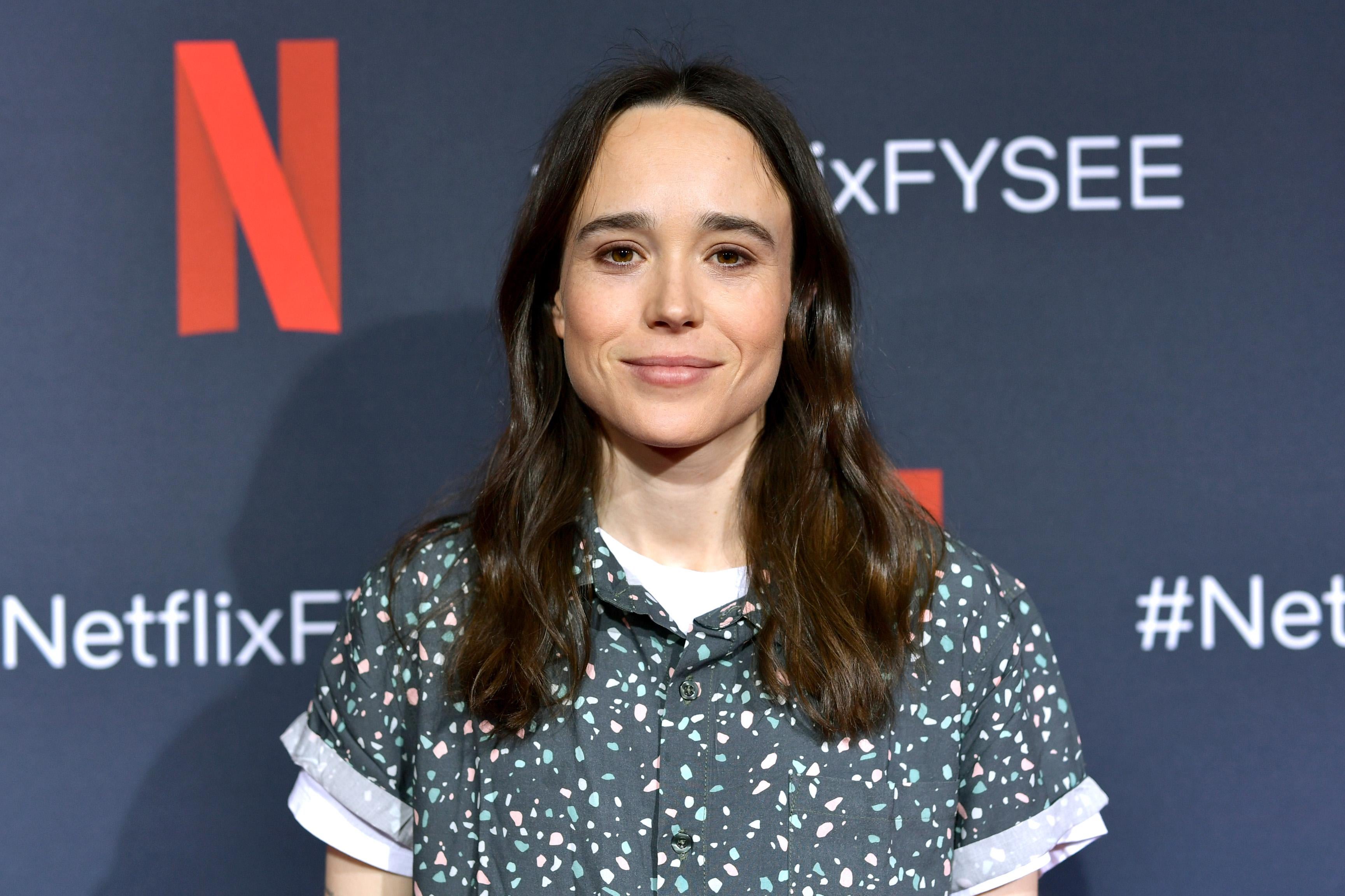 Elliot Page stands in front of a Netflix red carpet step and repeat.