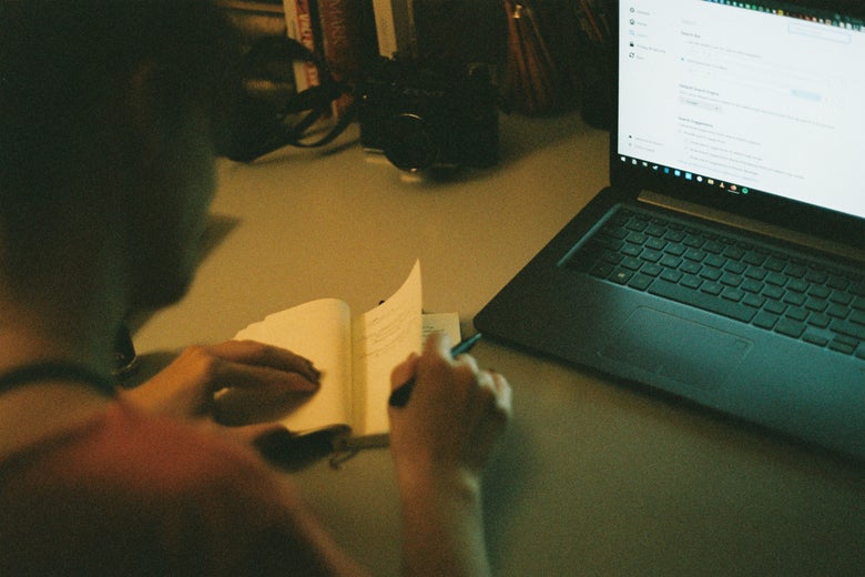 A person holds a notebook and pen in front of a laptop.