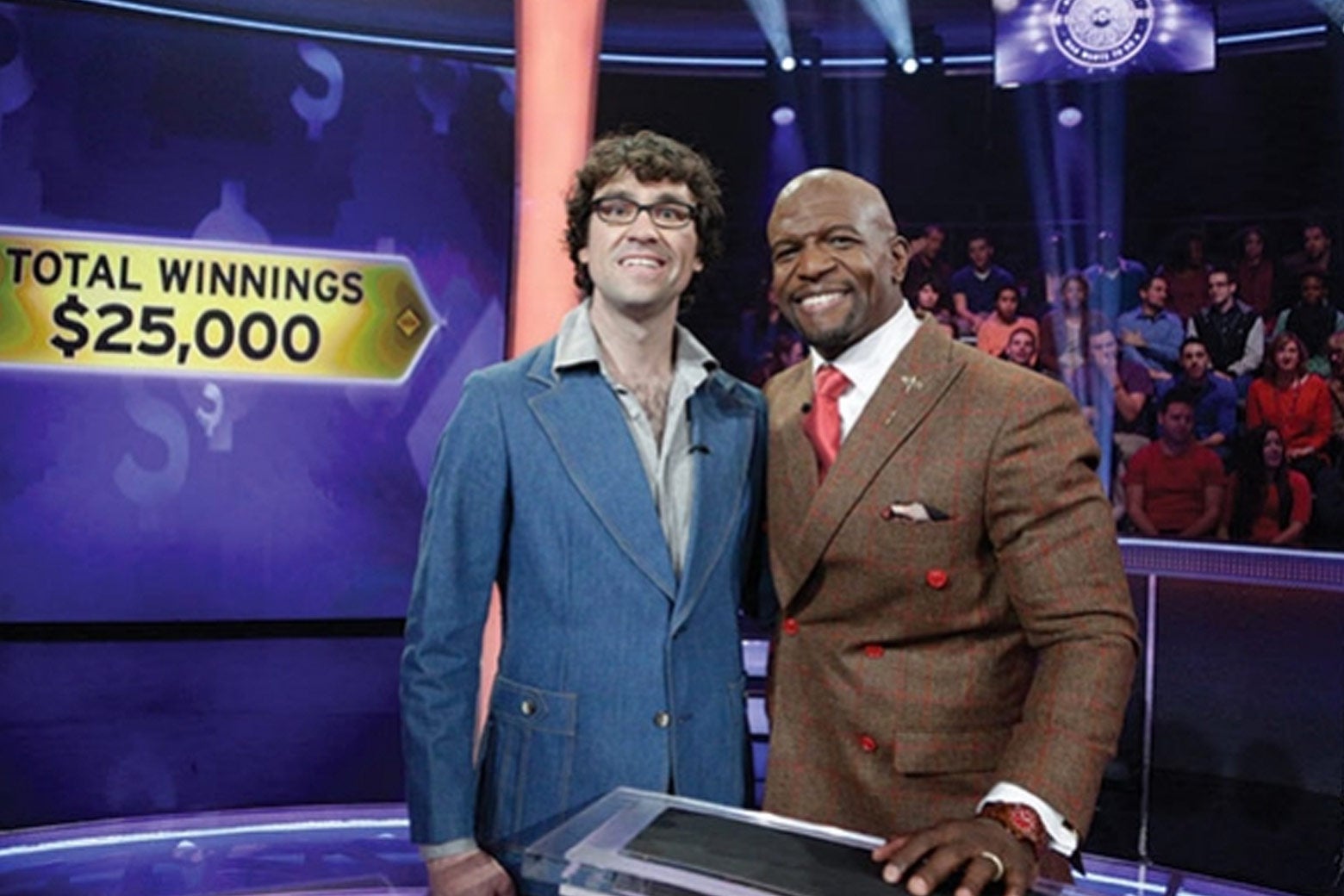 Terry Crews has his arm around Justin Peters on the set of Millionaire. Behind them, the studio audience and a screen that says "TOTAL WINNINGS $25,000."