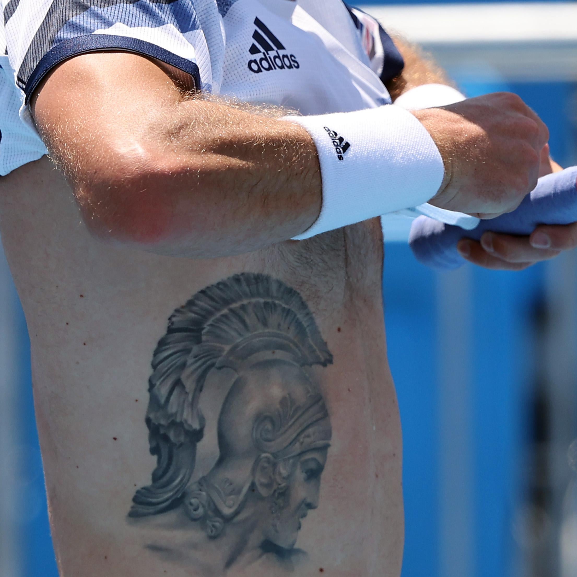 A man in Adidas wristband and holding a purple tennis-racket handle in his left hand lifts up his Adidas shirt, revealing the Achilles-in-helmet tattoo