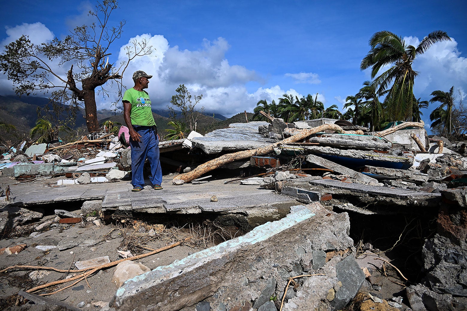 Мужчина в зеленой рубашке и бейсболке смотрит на разрушенное здание на Ямайке после урагана Мелисса.