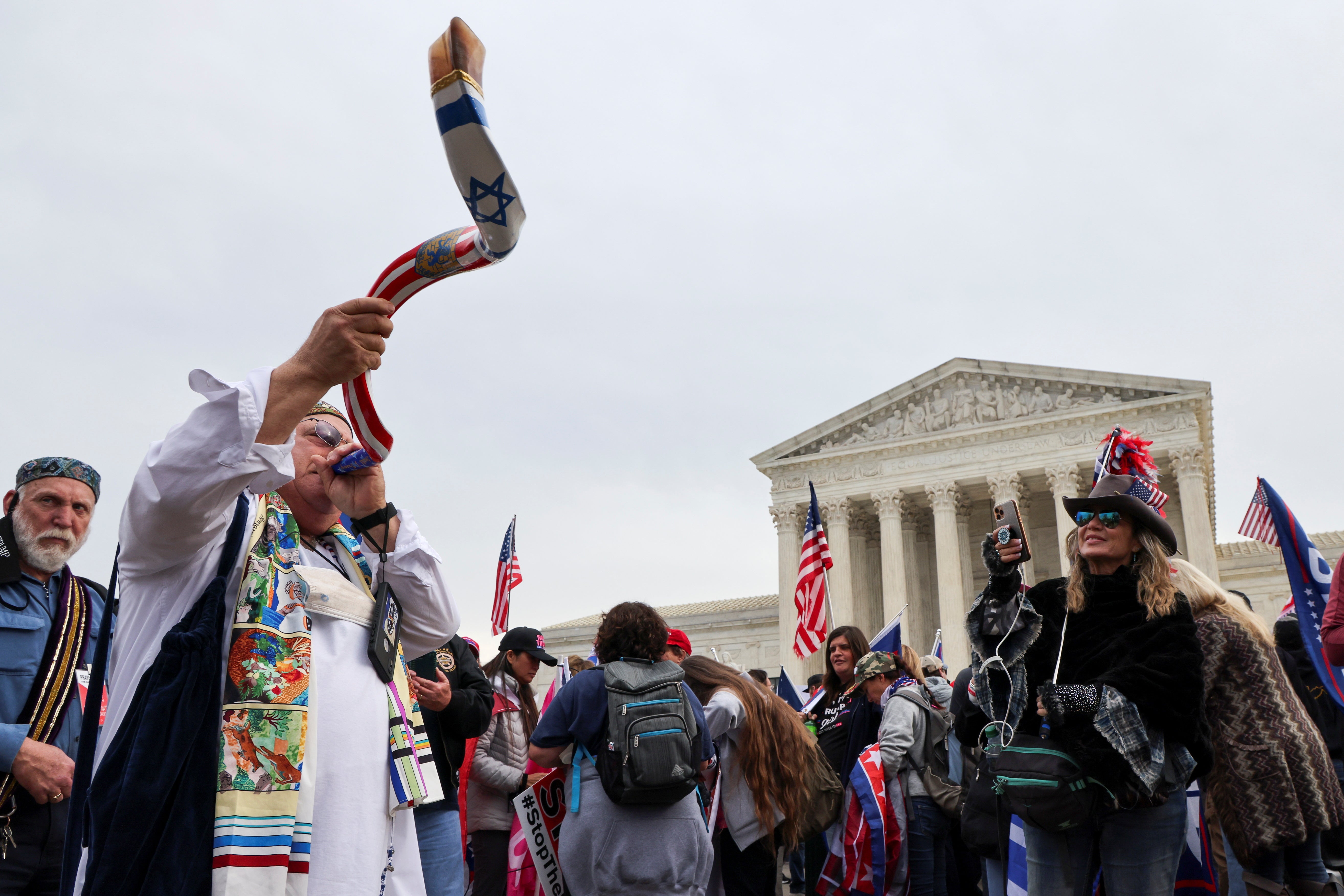 A man standing in a crowd in front of the Supreme Court building blows into a long, spiraling horn that is decorated with the American flag and Star of David. People in the crowd are wearing red, white, and blue and holding Stop the Steal signs. 