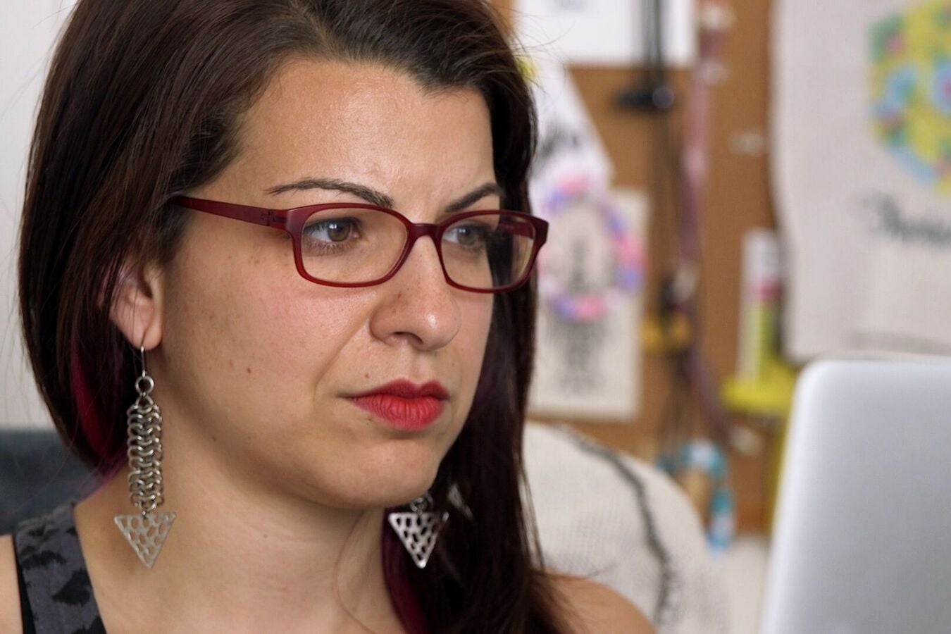 Feminist Frequency founder Anita Sarkeesian, a woman with white skin and dark brown hair wearing magenta glasses, looks at her computer screen.