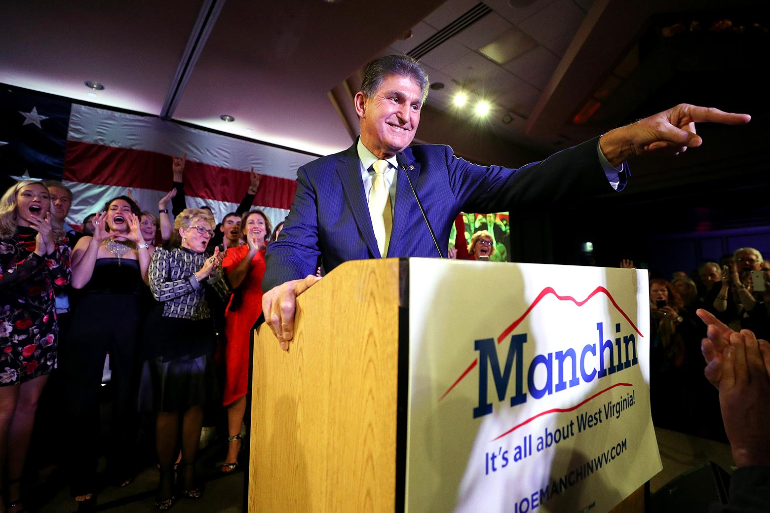 Joe Manchin smiles and points into the crowd as he stands behind a podium that says, "Manchin: It's all about West Virginia."