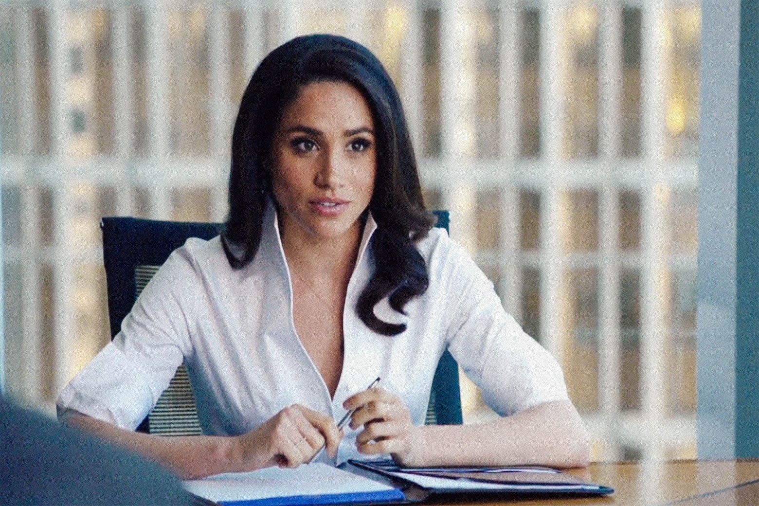 Meghan Markle sits at a desk with an open notebook in front of her. She's wearing a white blouse.