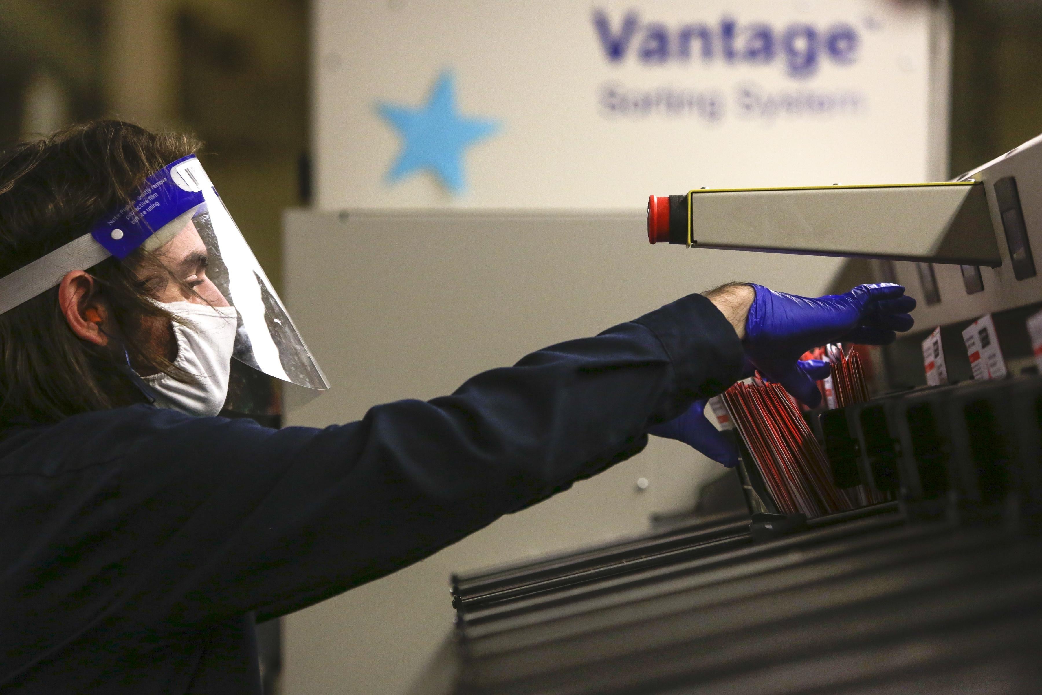 A man wearing a face shield and mask takes ballots from a sorting machine.