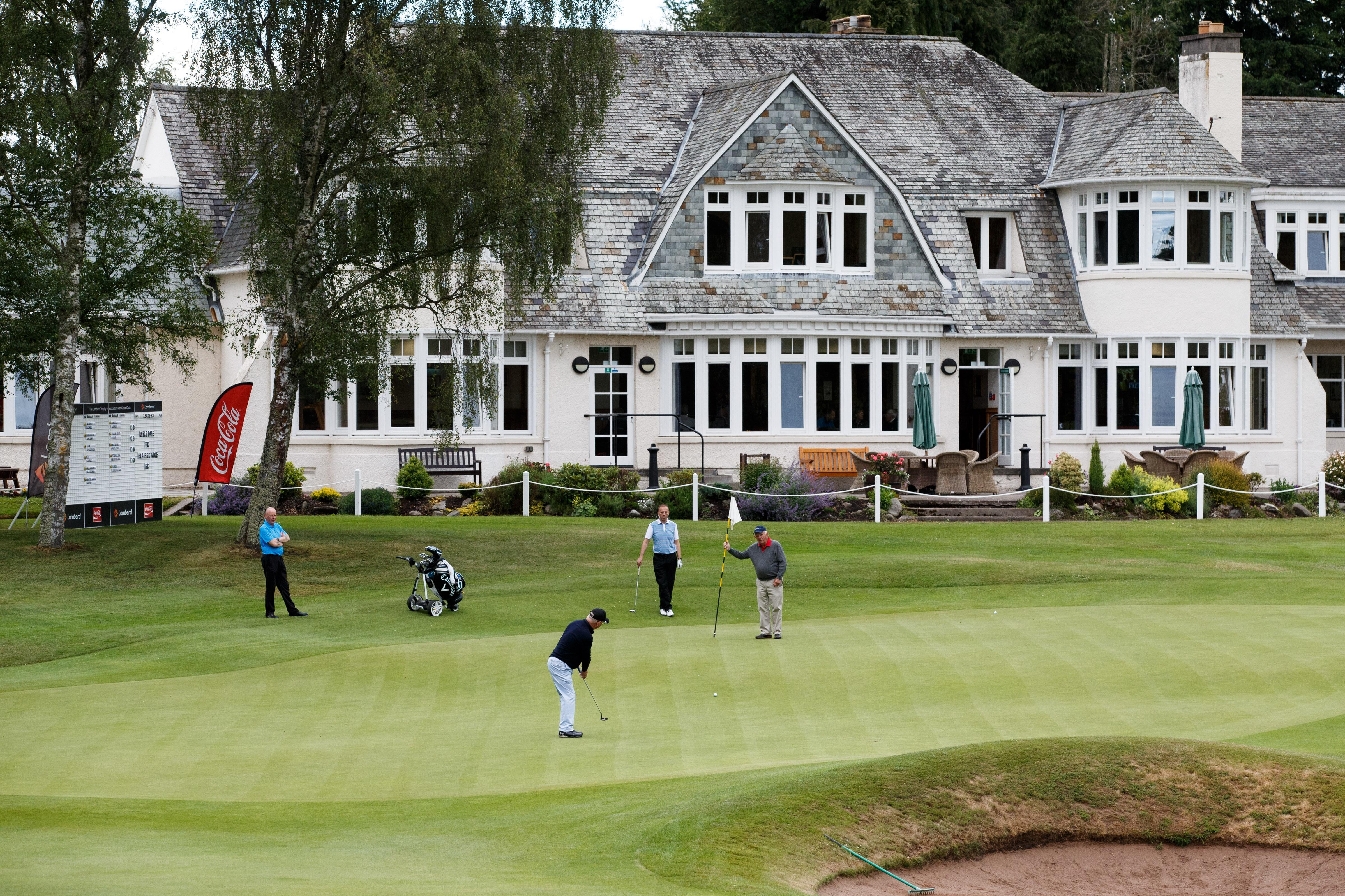 Four men stand on a putting green, a large club house stands in the background.