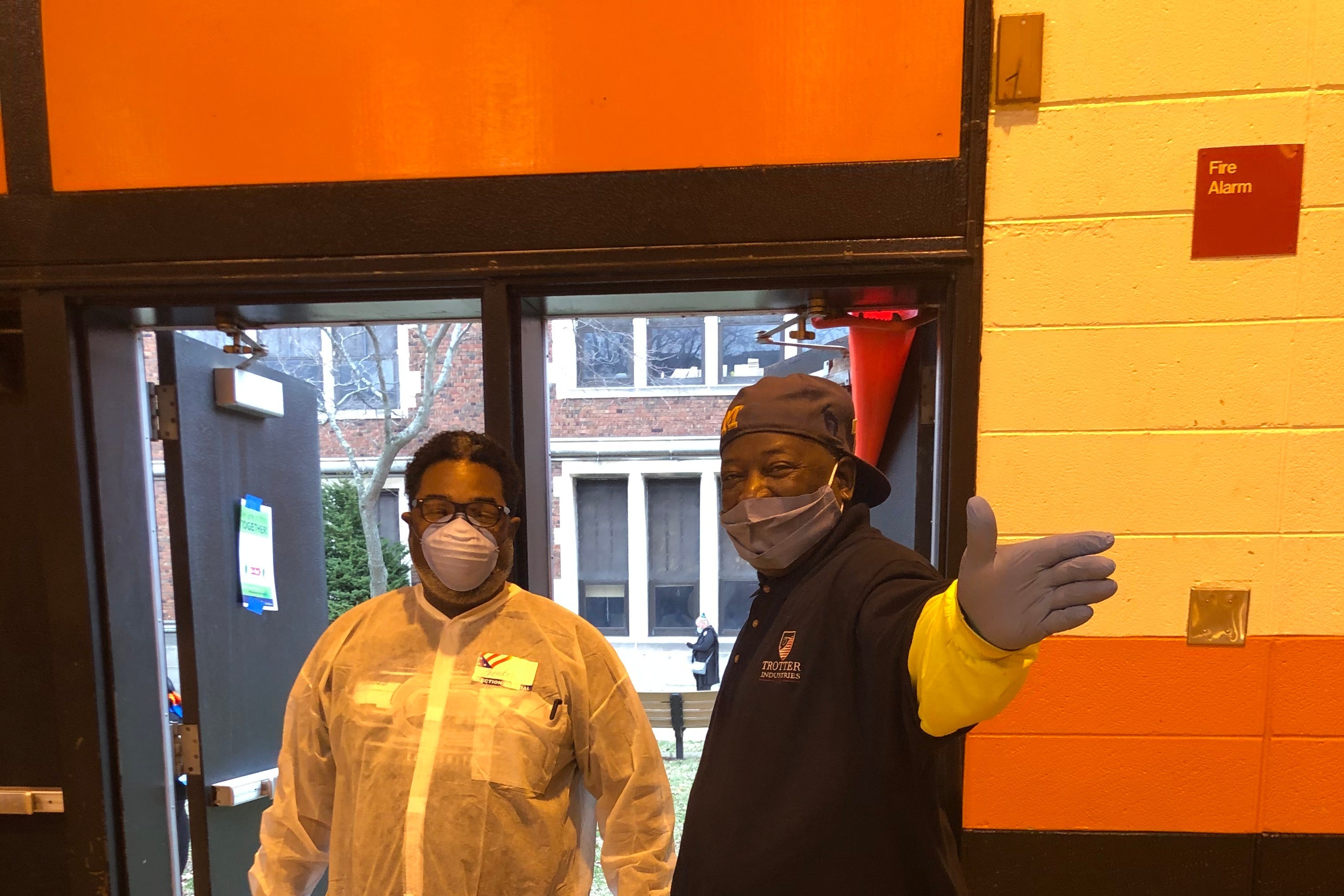 Poll workers in gowns, gloves, and masks stand by a door