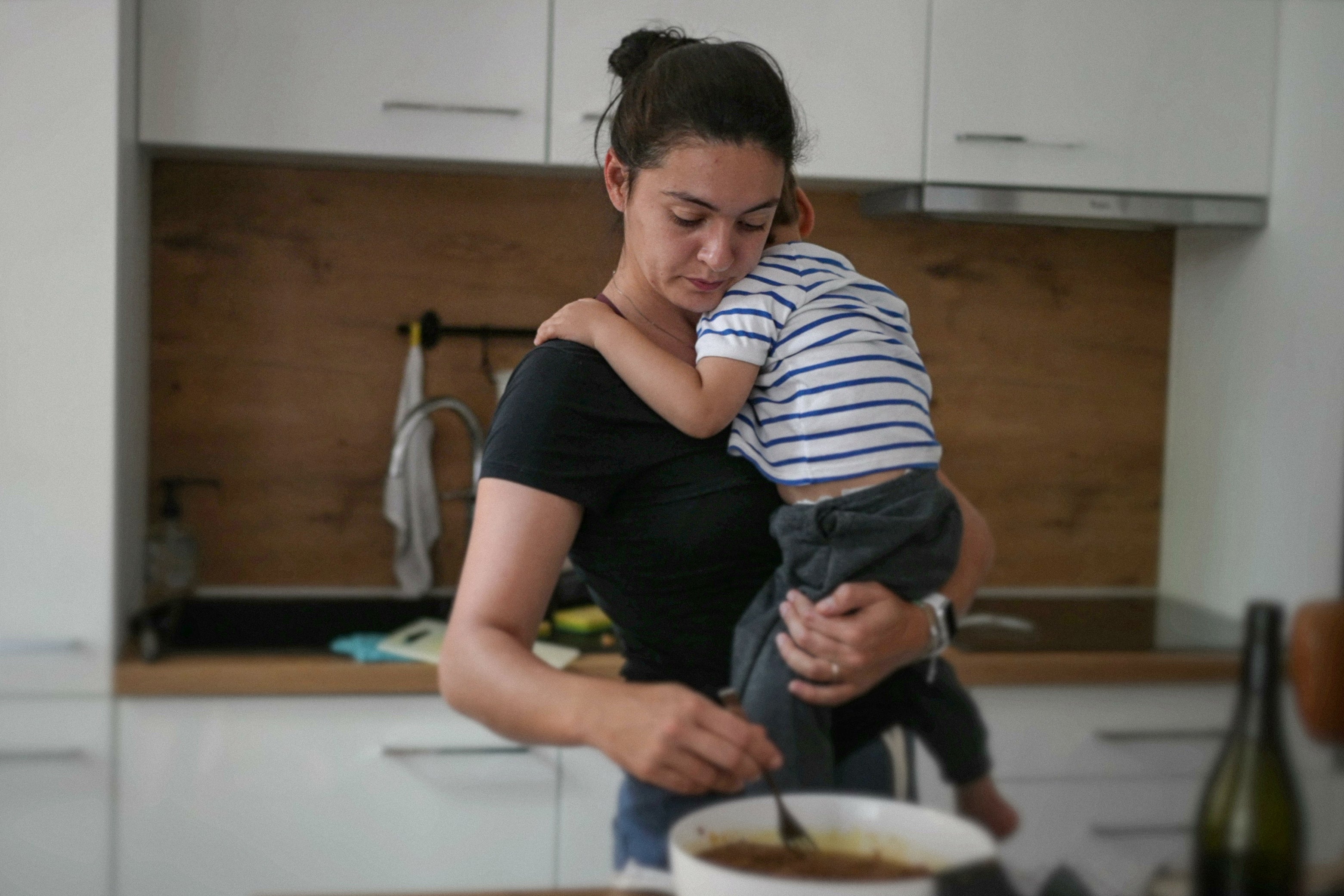 A woman stirs something on a stove while also holding a toddler. 