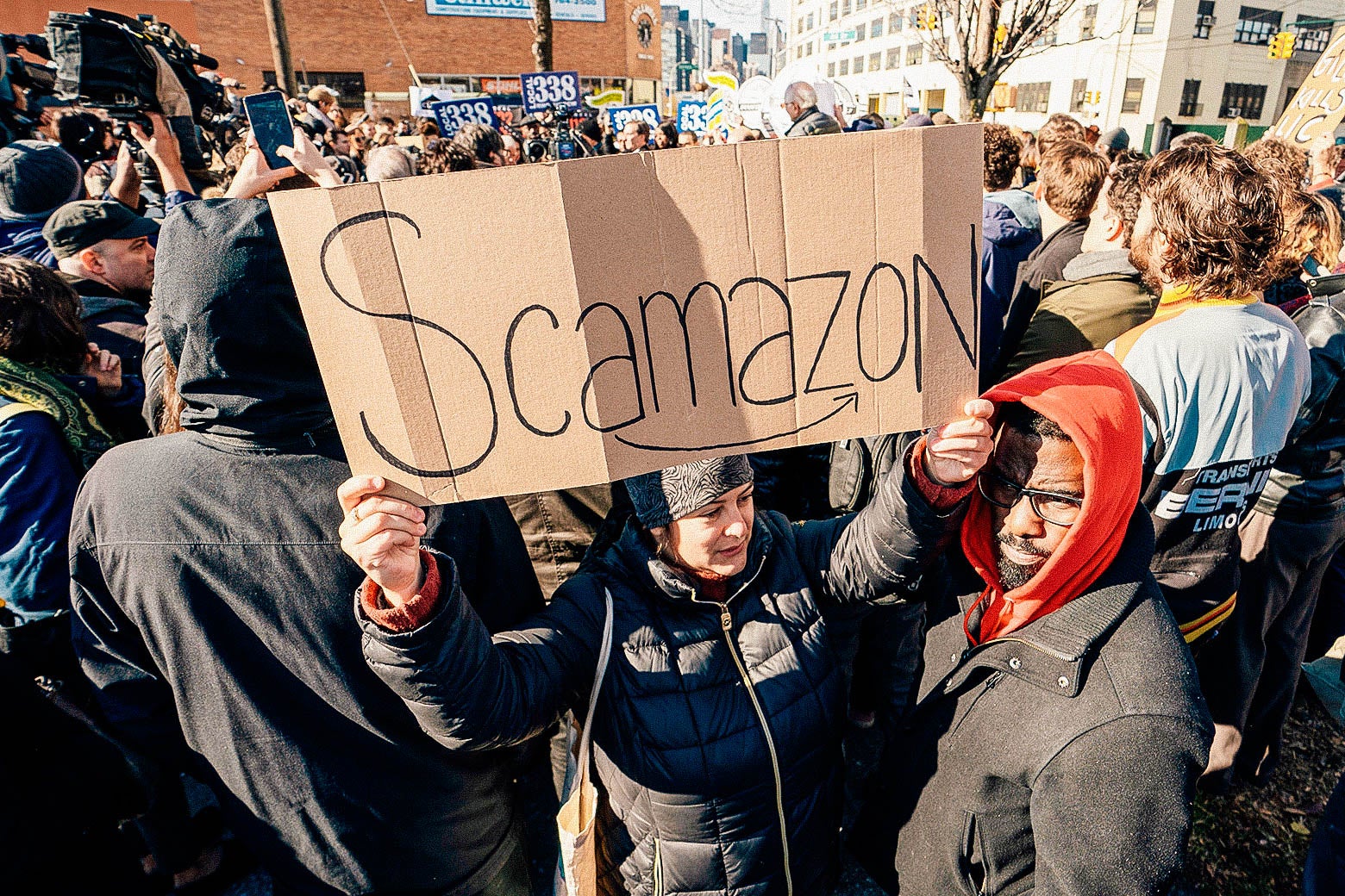 A crowd of anti-Amazon protesters, one holding a "Scamazon" sign.