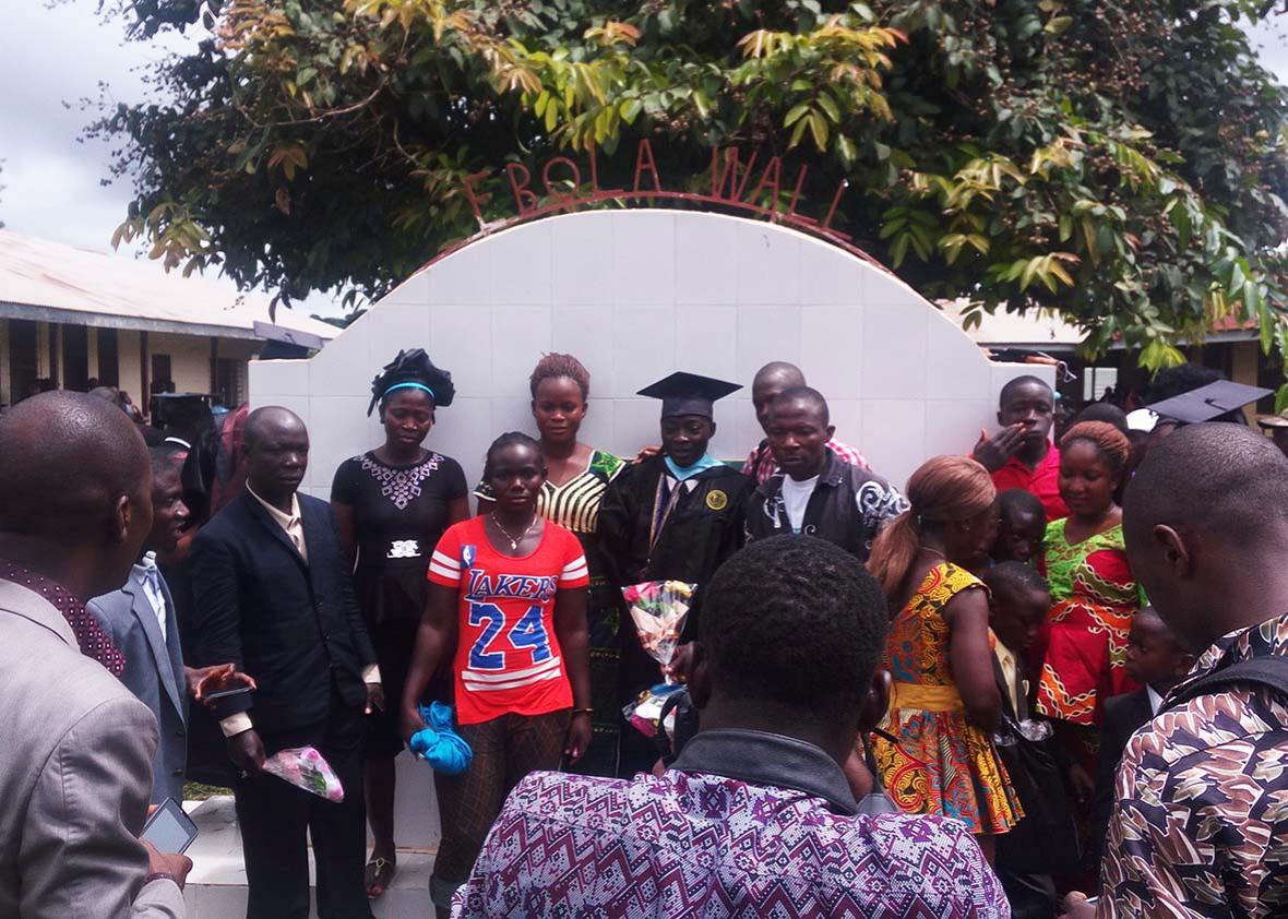 A graduate lines up in front of the Ebola Wall with family and friends after graduation for photos. The Ebola Wall was built to honor the 19 CU faculty and students who dies from Ebola.  Most were grads of the schools nursing program, one of the few in the country. Classrooms can be seen to the left and right.