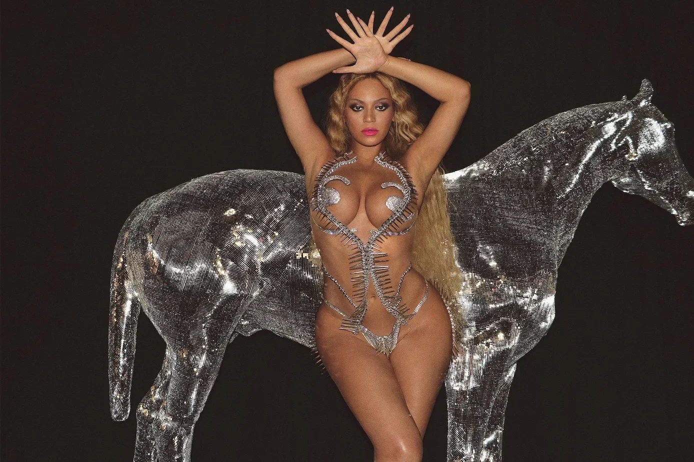 The singer poses in front of a black background and what looks like a horse made out of a disco ball. Her hands are over her head, and she wears a sort of barely-there metal-and-jewels bikini.