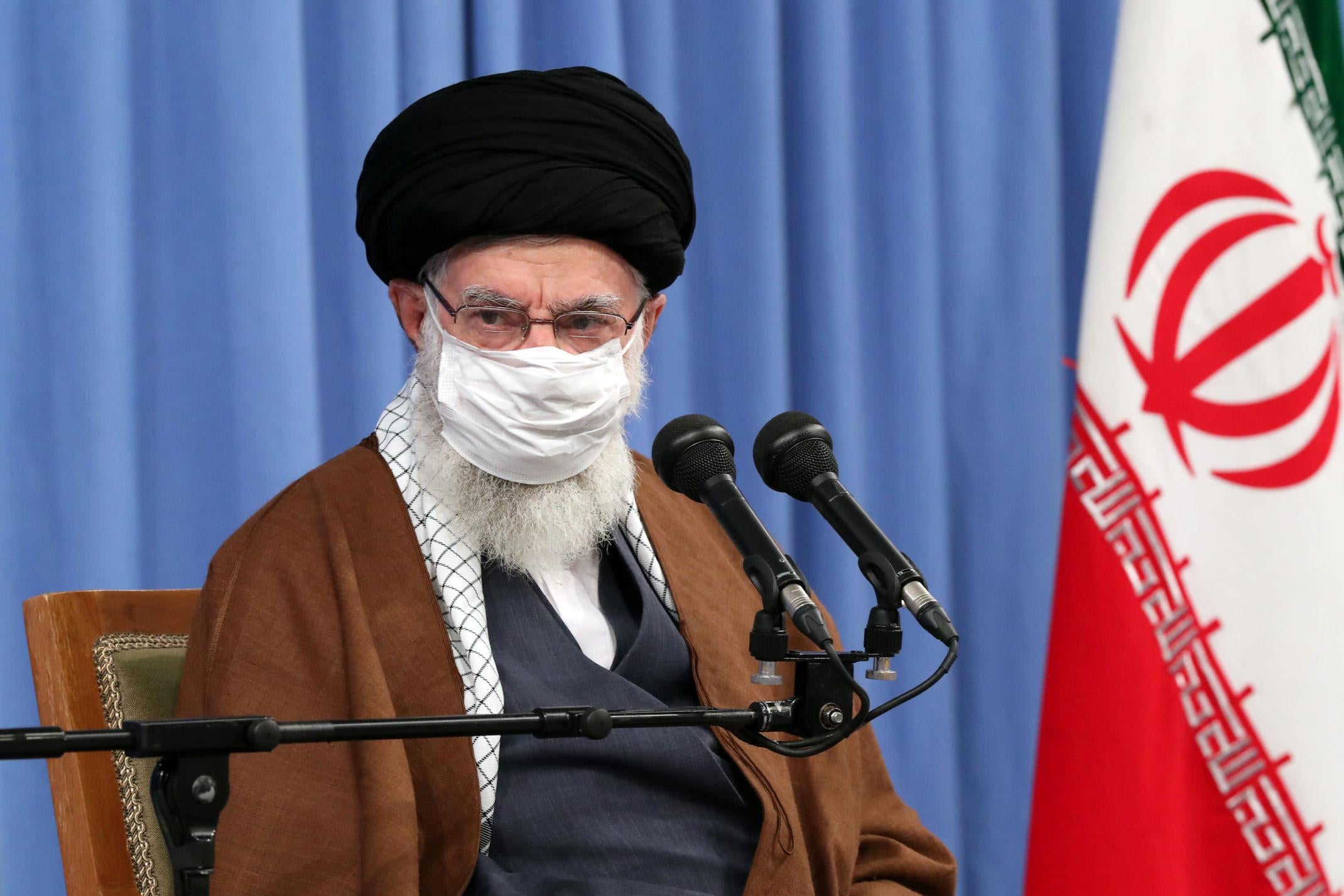 Khamenei, wearing a mask, sits at a mic with an Iranian flag to the side