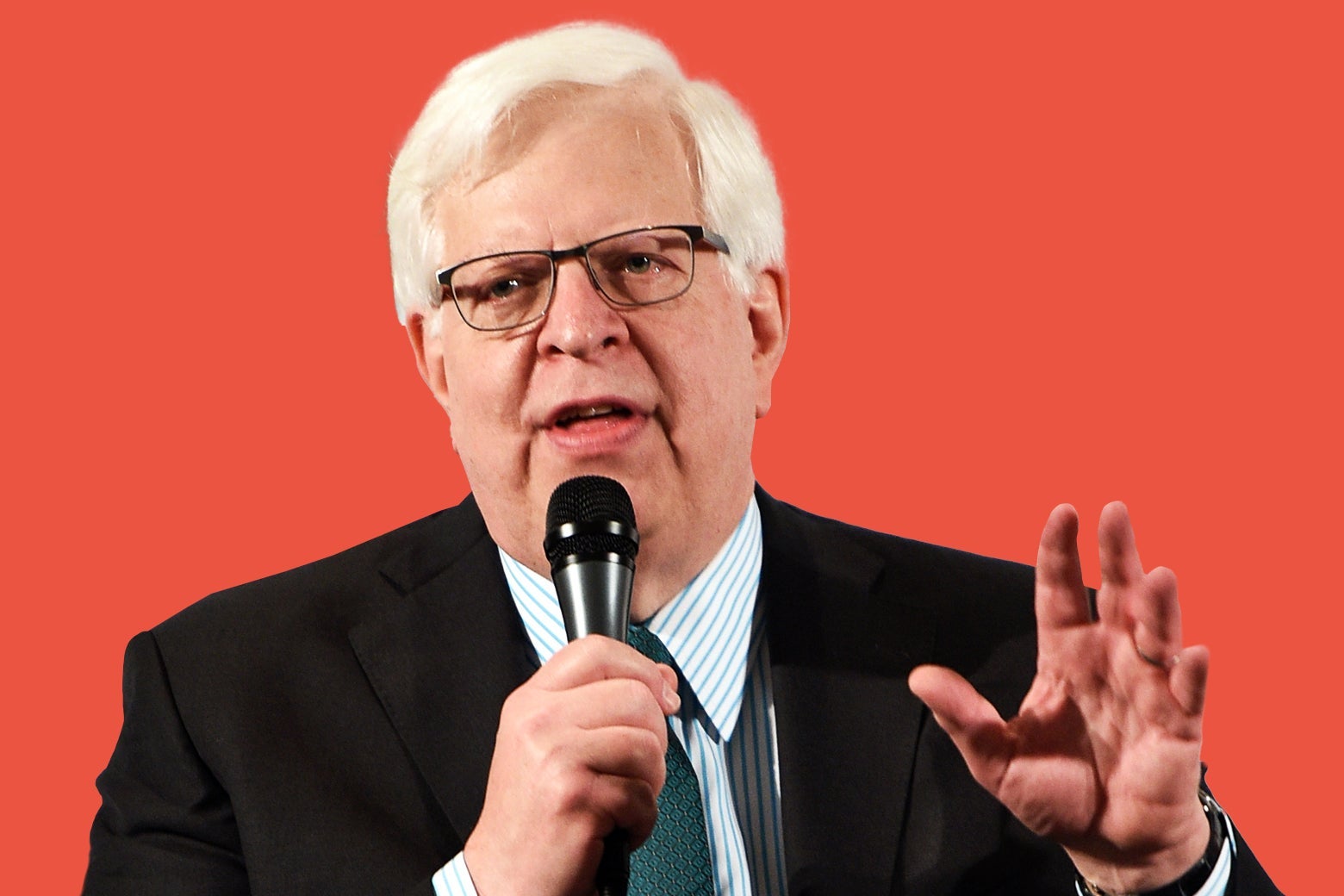 A white man with white hair and glasses holds up one hand while holding a microphone to his mouth with the other hand.
