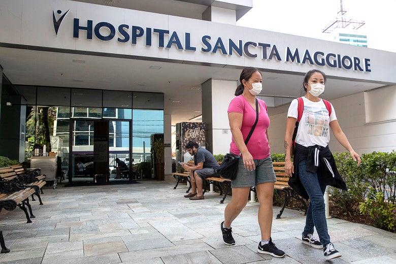 Two women, wearing face masks, walk out of a hospital. Someone sits on a bench in the background.
